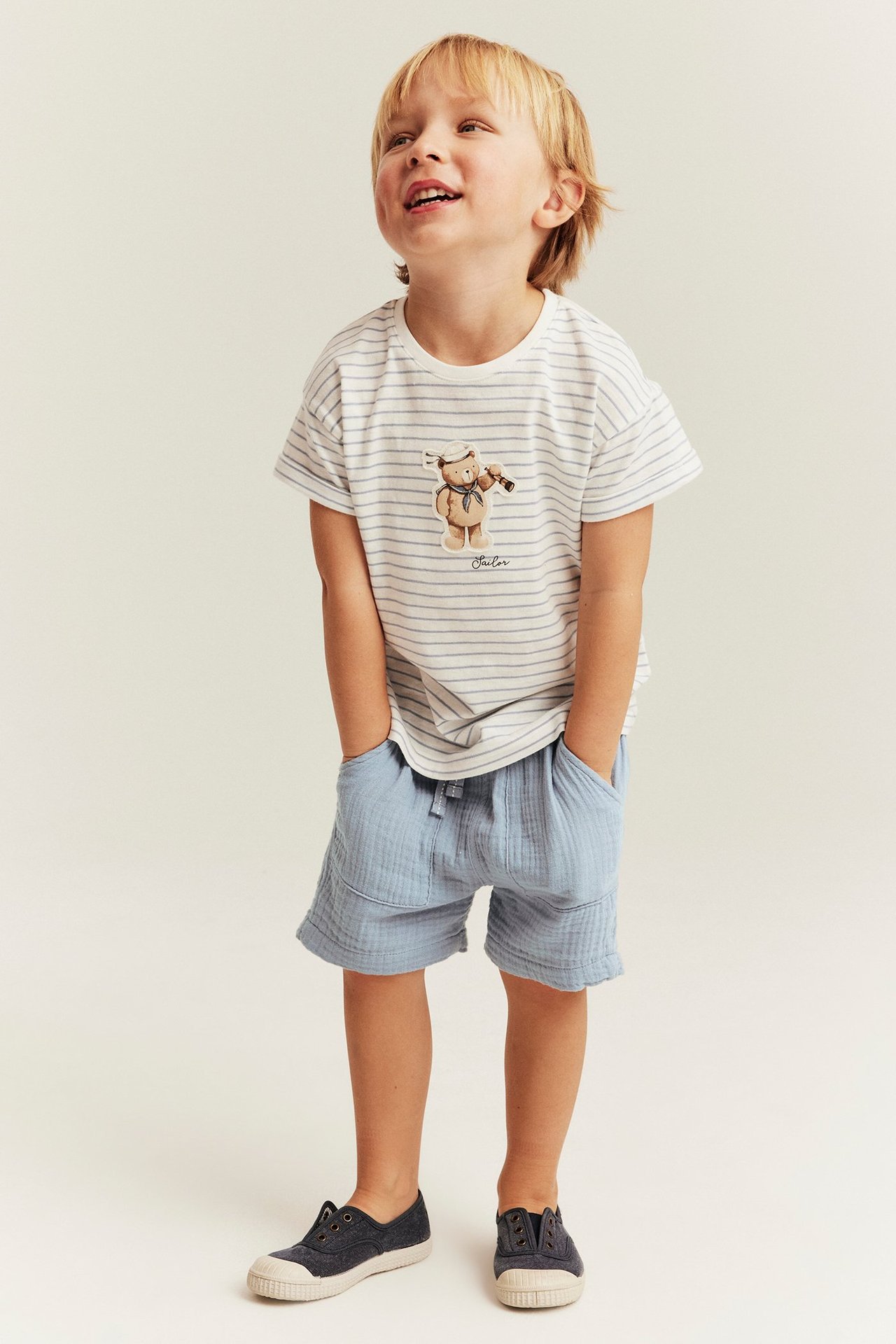 Vorderansicht: Junge in blauen Baumwollshorts mit Taschen, gestreiftem T-Shirt, dunkelblauen Schuhen.