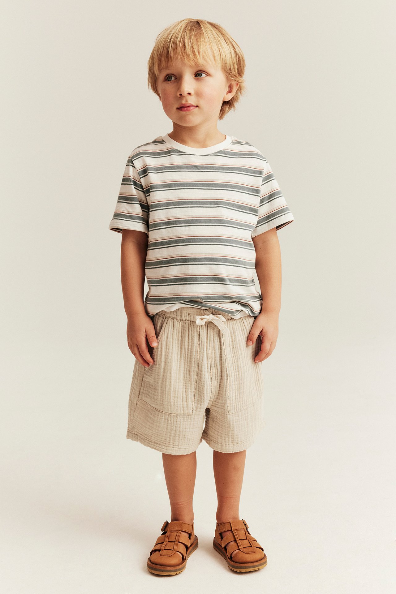 Front view of a kid wearing offwhite woven cotton shorts with drawstring, a striped T-shirt, and brown sandals.