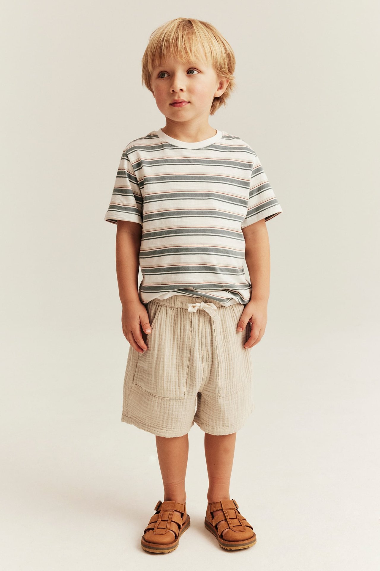 Vorderansicht Kind in beigefarbenen Baumwollshorts mit Kordelzug, gestreiftem T-Shirt und braunen Sandalen.