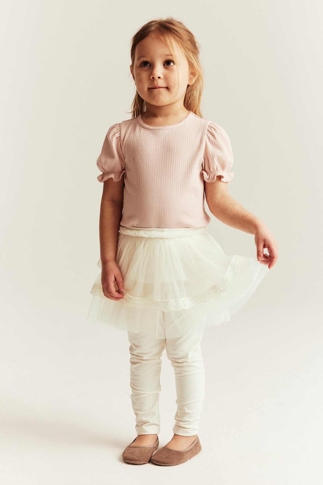 Front view: Kid wears offwhite cotton leggings with attached three-ruffle tulle skirt, pink ribbed top, and brown shoes.