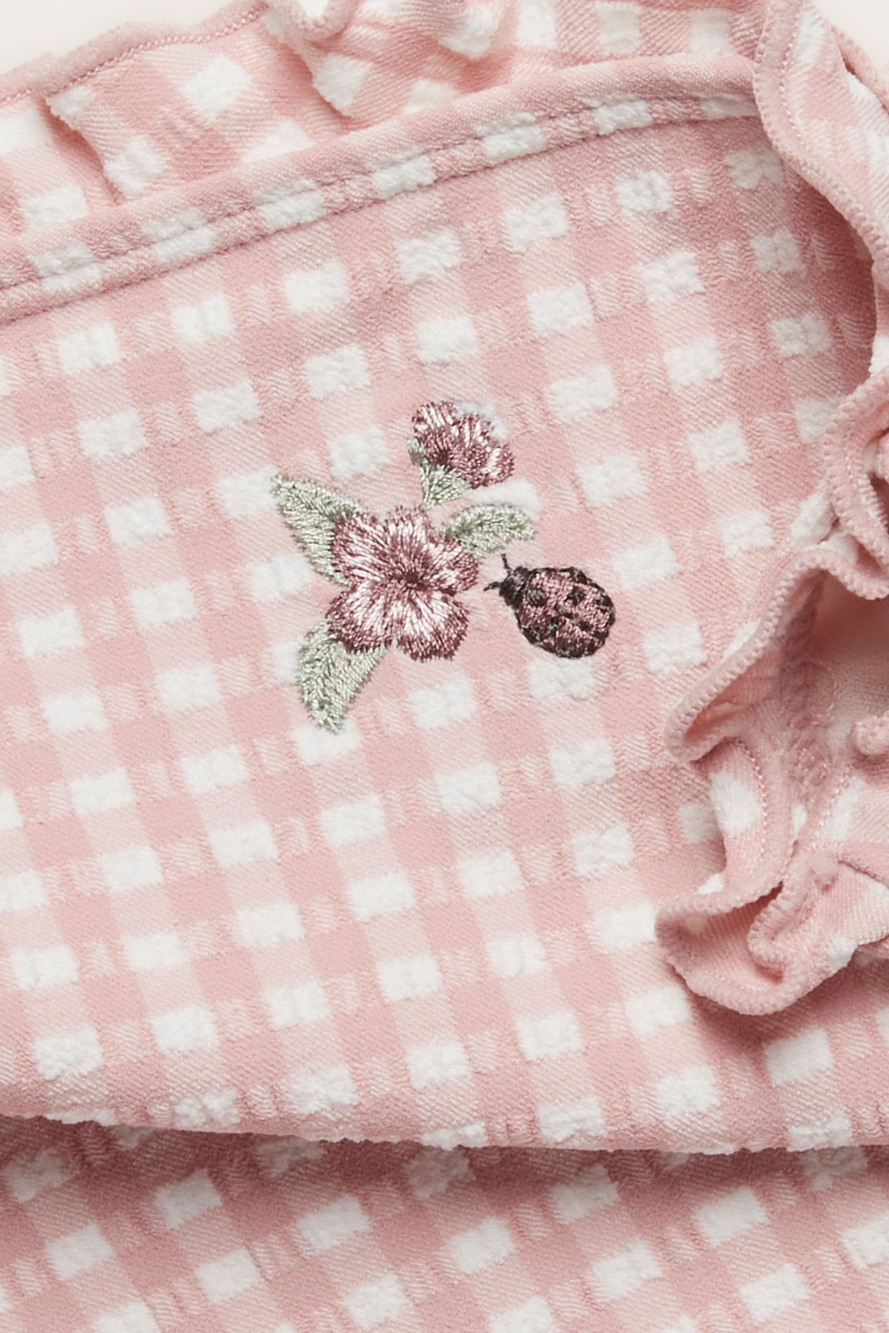 Close-up of a pink and white checked seersucker swimsuit with embroidered flower, ladybug, and ruffles.