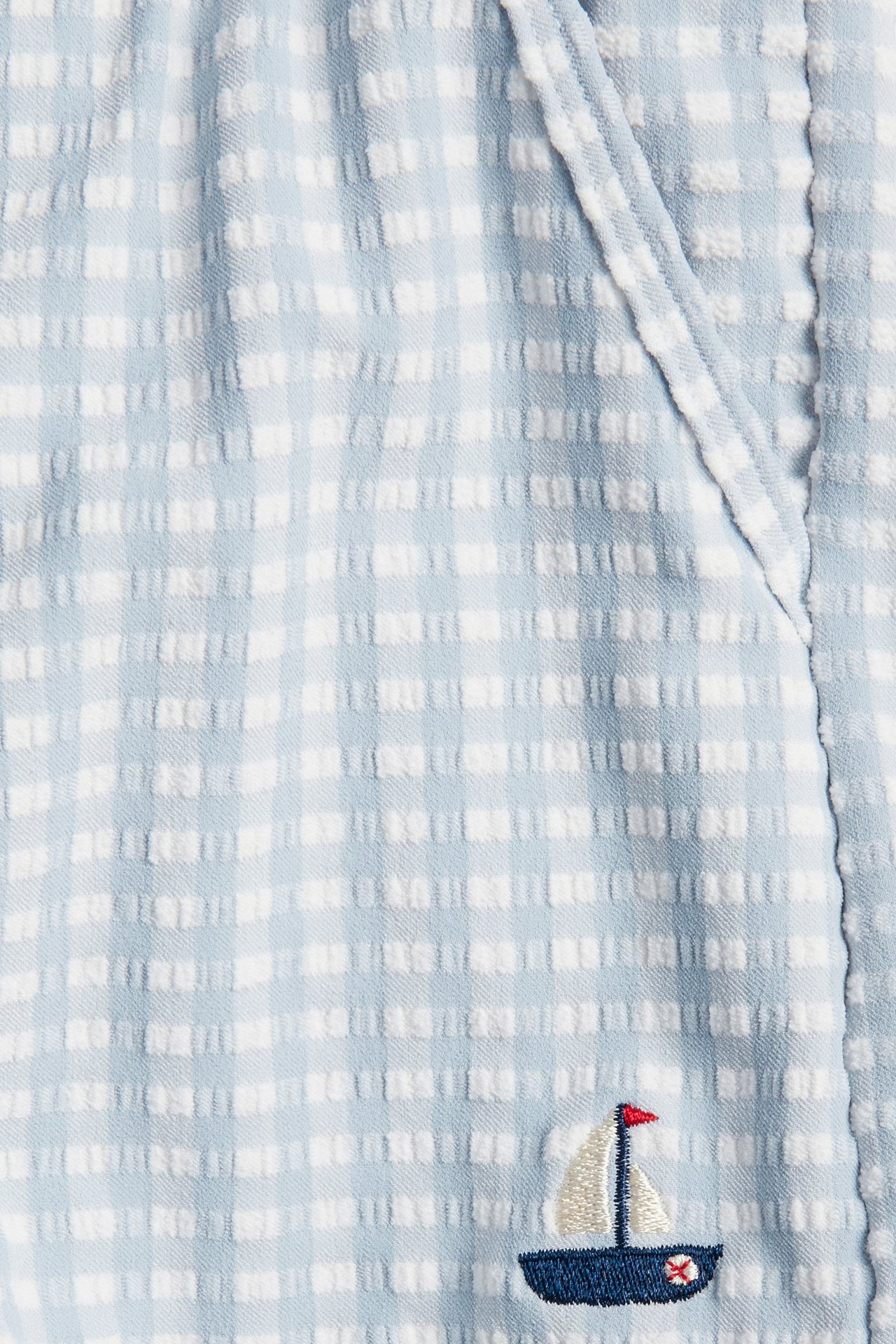 Close-up of light blue and white checked seersucker swim shorts with an embroidered boat.