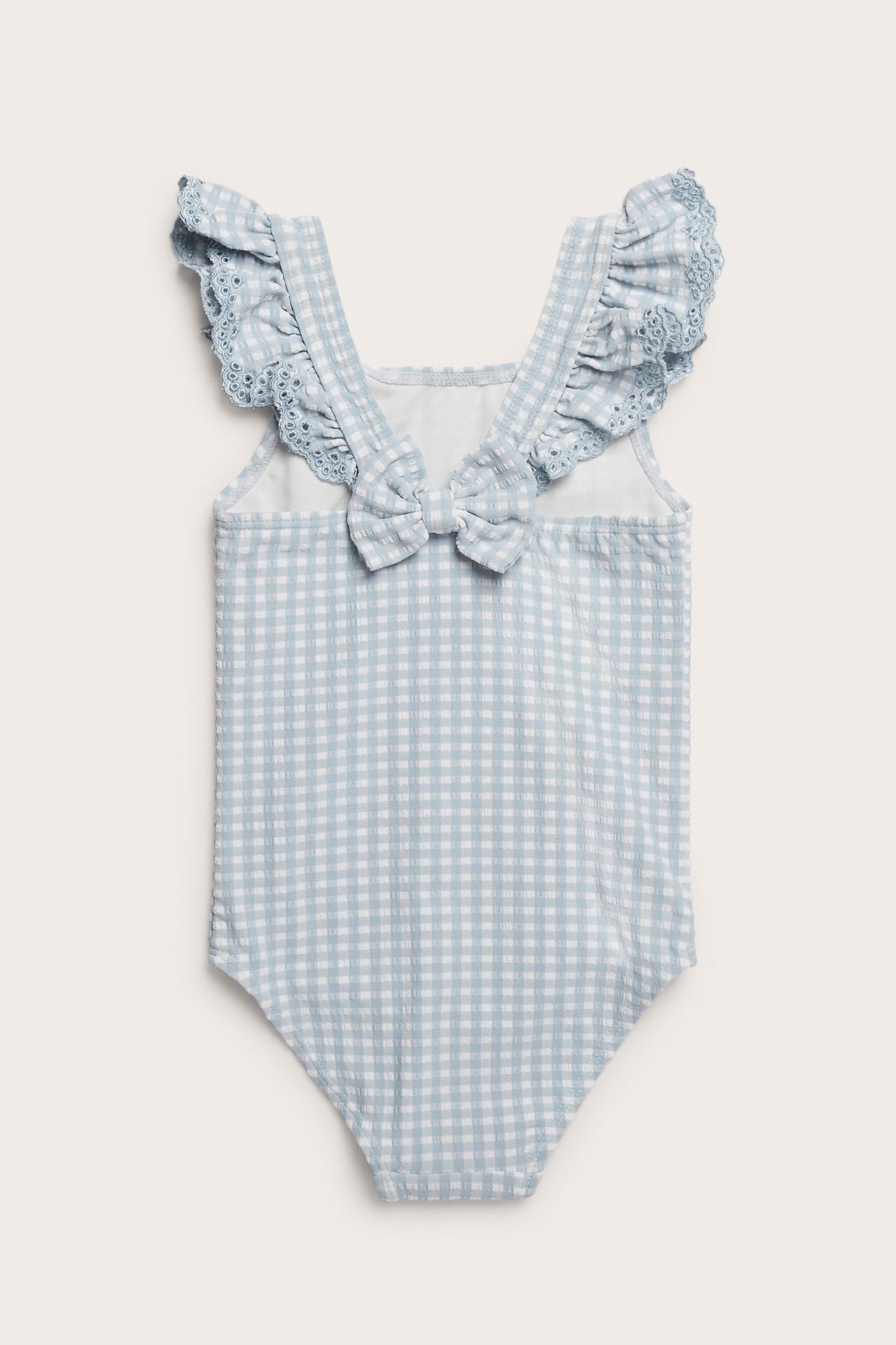 Back view of a kid's light blue and white gingham seersucker swimsuit with ruffled, embroidered straps and a back bow.