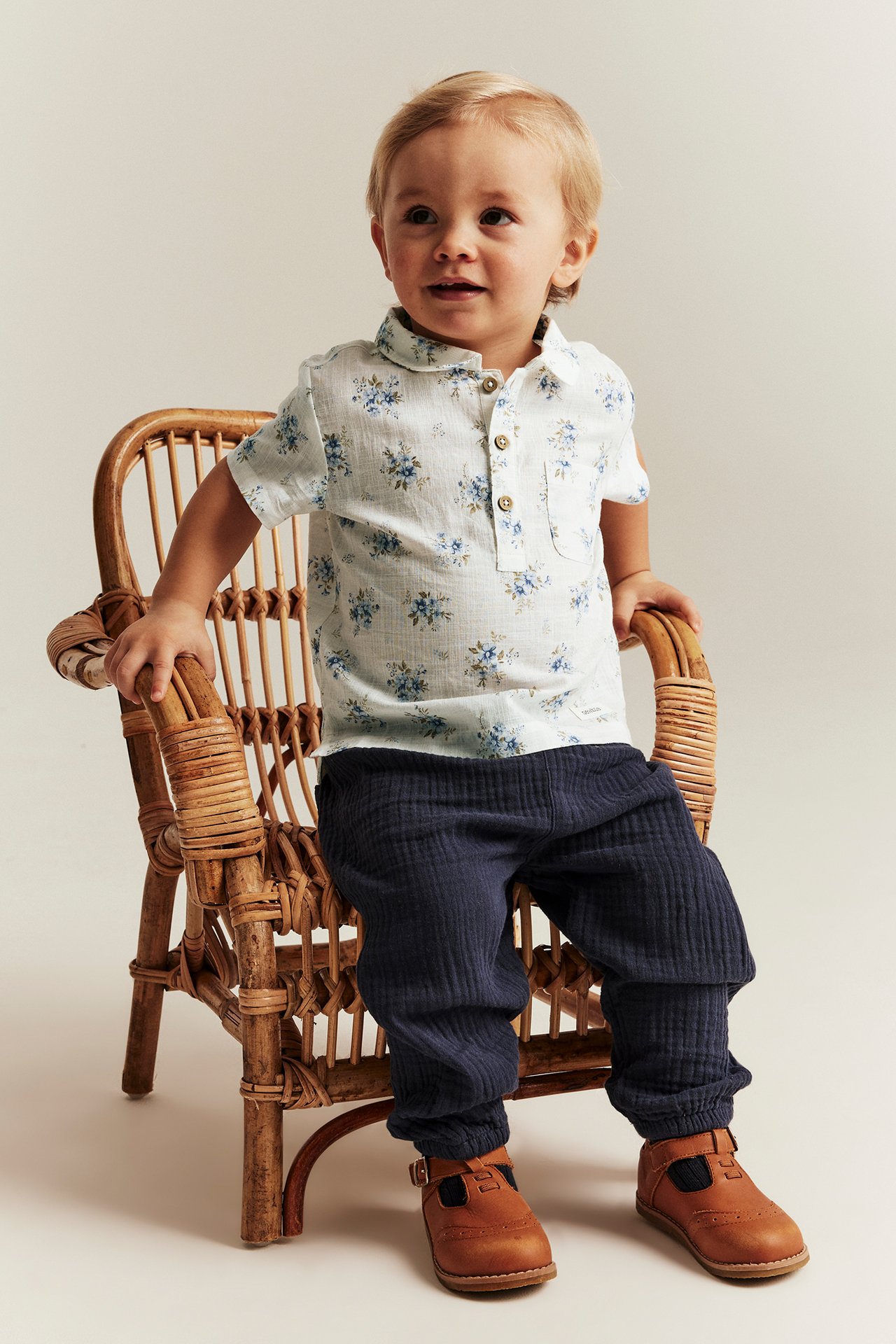 Kid in an off-white floral short-sleeved cotton shirt, dark blue textured pants, and brown buckle shoes.
