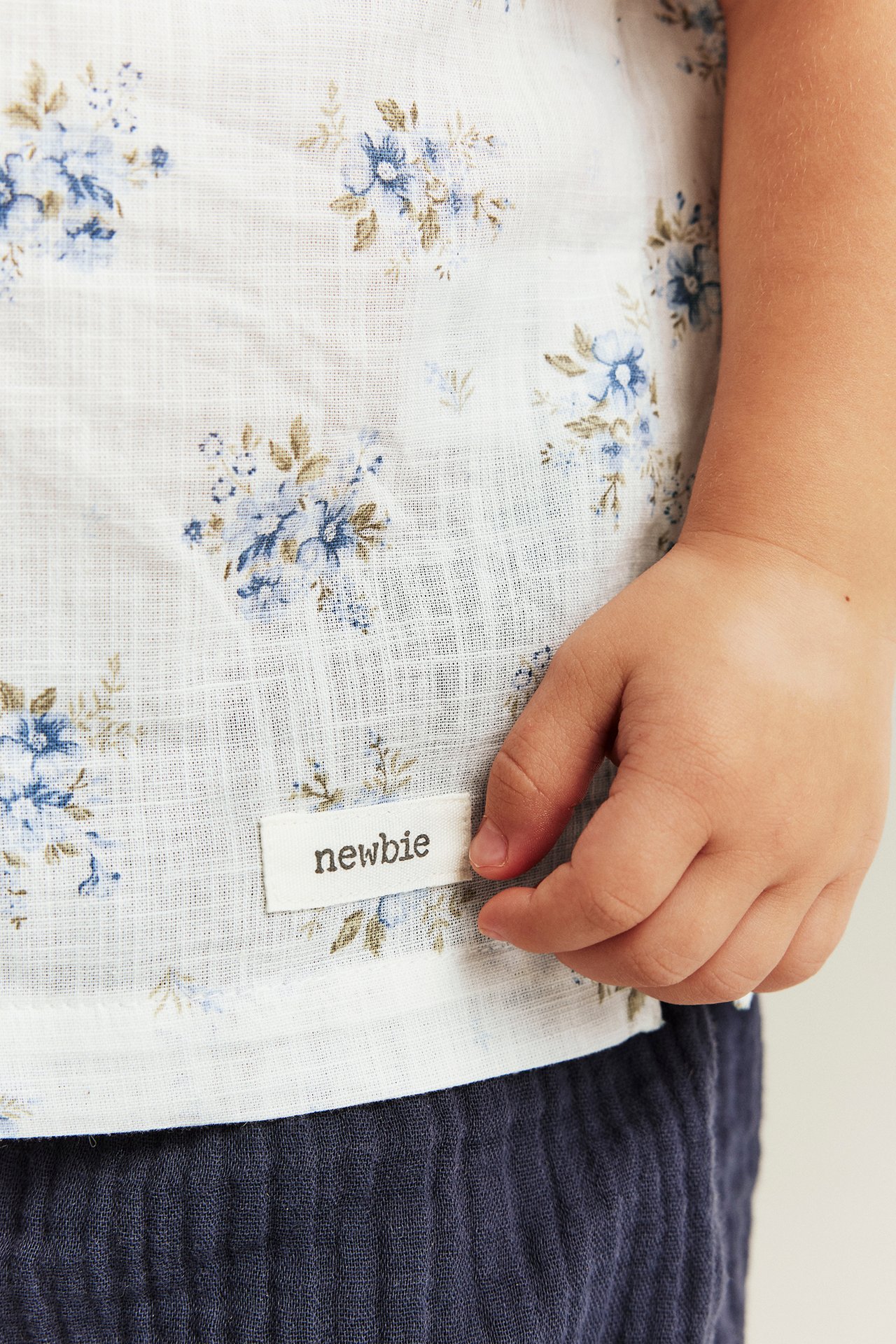 Close-up of a kid's offwhite cotton shirt with blue floral pattern, held by a hand over dark blue pants.