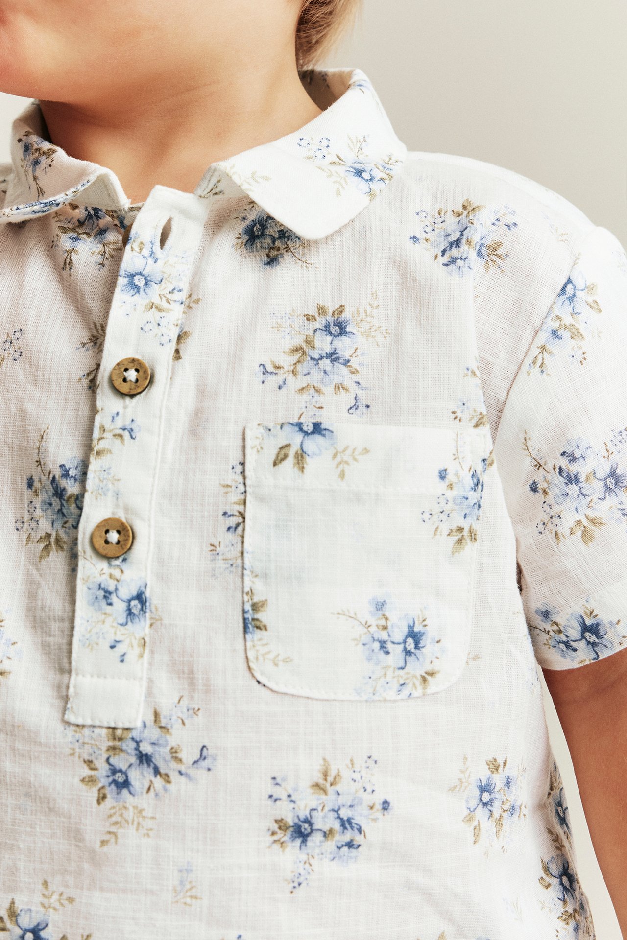 Close-up front: kid wears off-white short-sleeved cotton floral shirt with collar, buttons, and pocket.