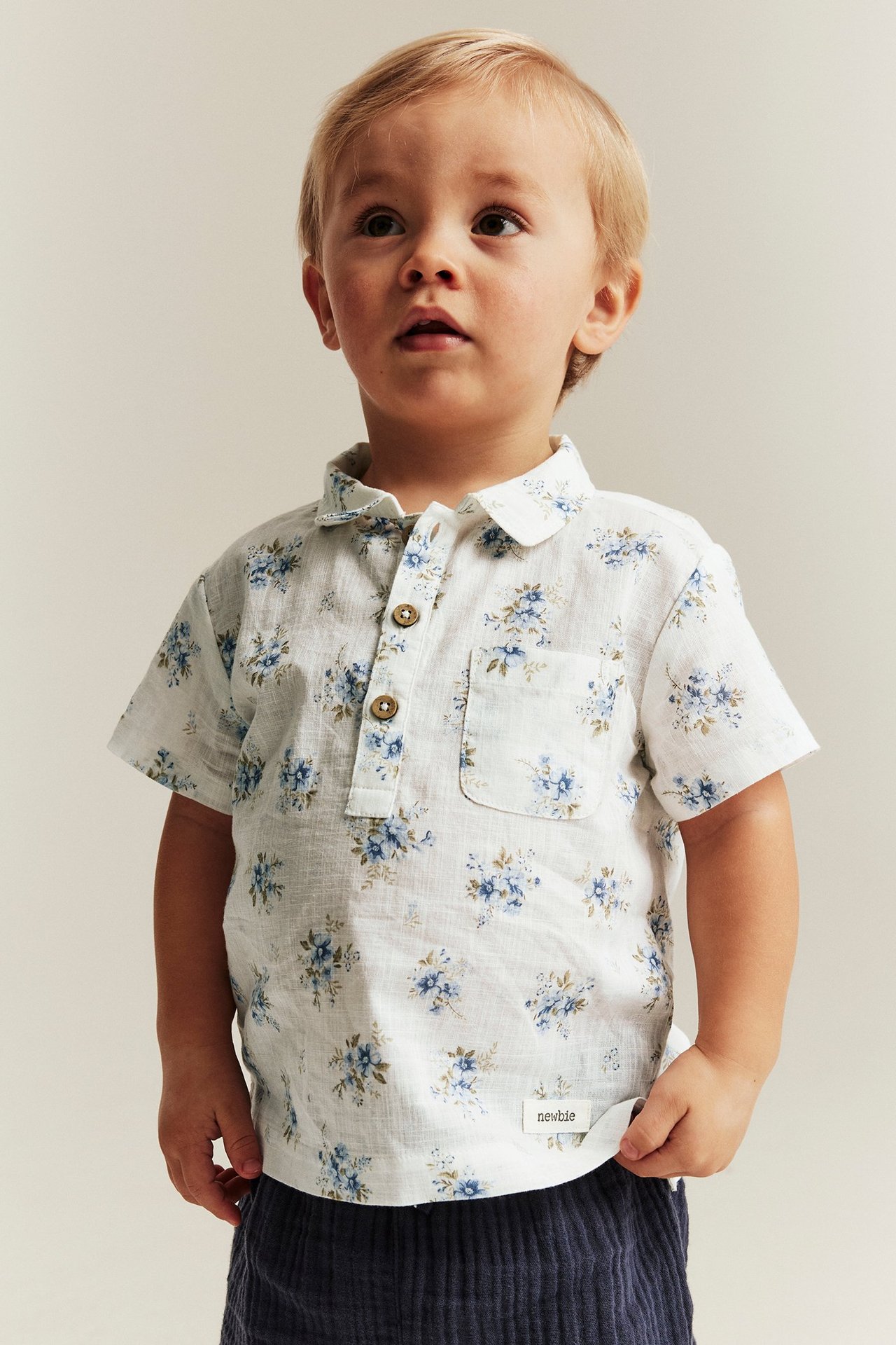 Front view of a kid wearing an off-white cotton short-sleeved floral shirt with blue flowers and dark blue trousers.