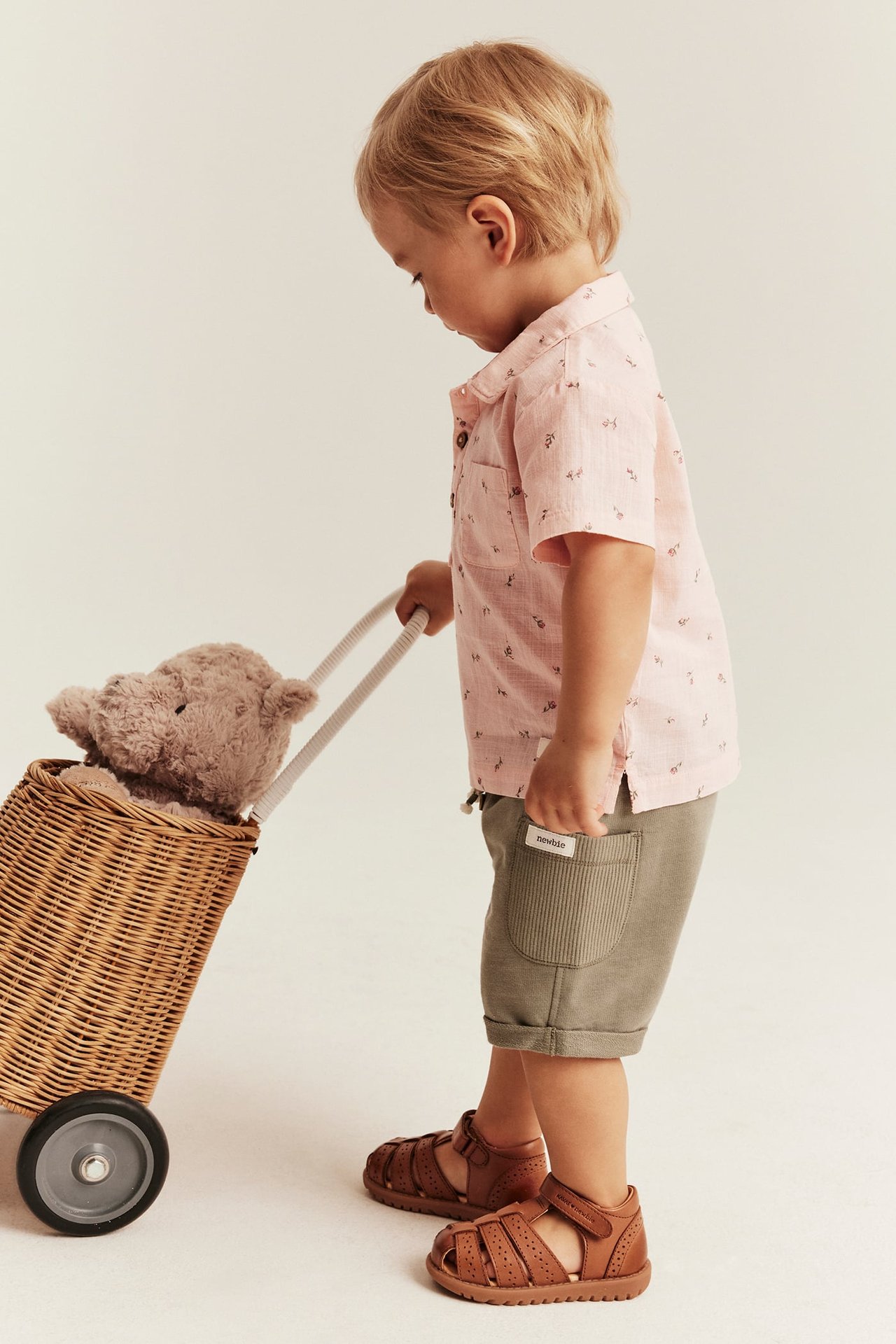 Kind trägt rosa geblümtes Kurzarmhemd, grüne Shorts und braune Sandalen, zieht einen Korb.
