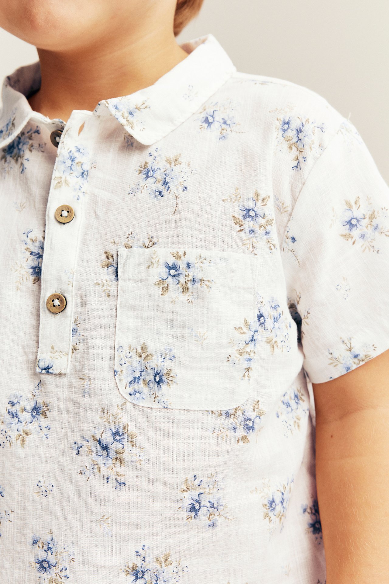 Nahaufnahme: Kinderhemd aus cremeweißer Baumwolle mit kurzen Ärmeln, blauem Blumenmuster, Kragen und Brusttasche.