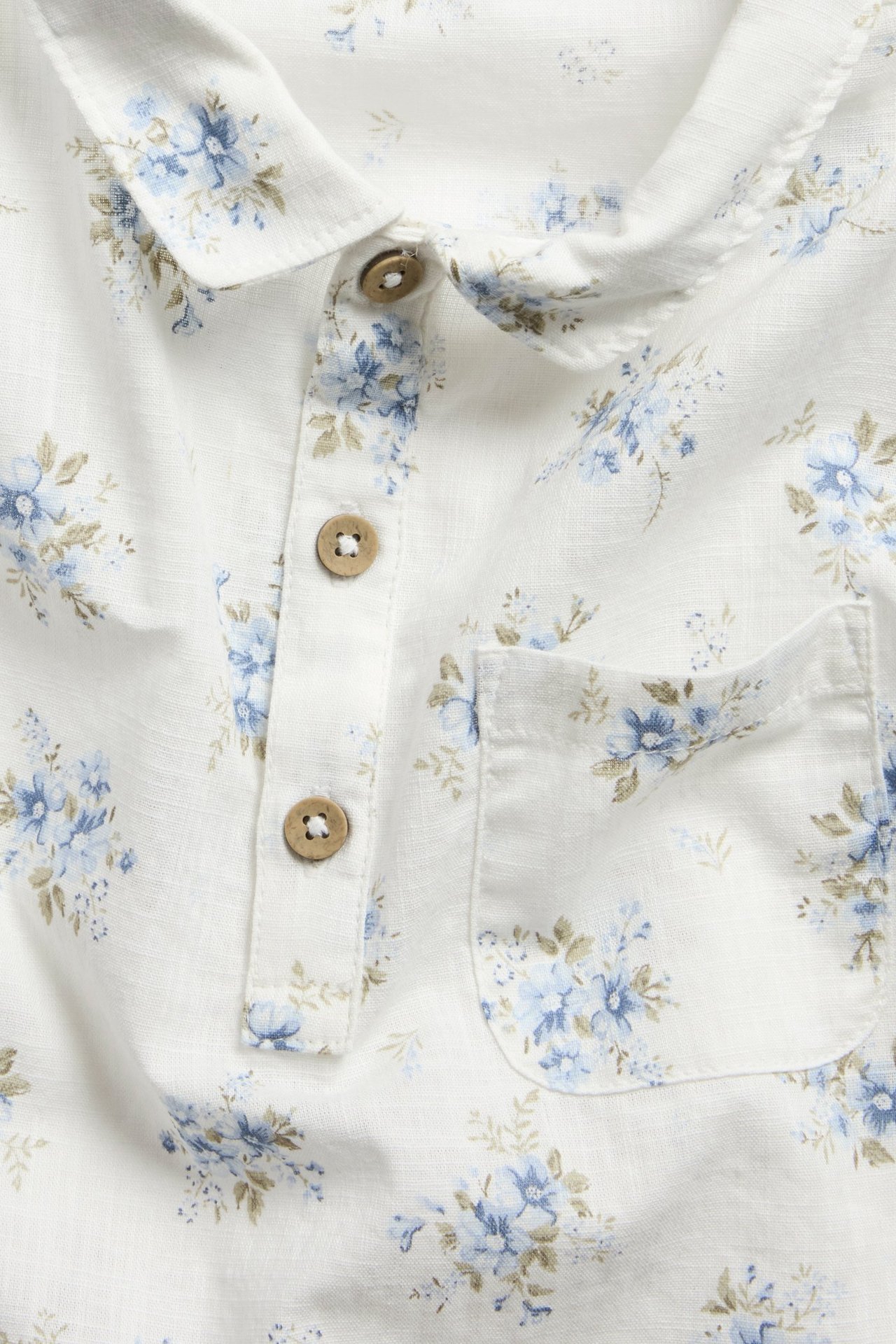 Nahaufnahme vorne: Cremeweißes kurzärmeliges Baumwollhemd mit blauem Blumenmuster, Kragen, Knopfleiste und Brusttasche.