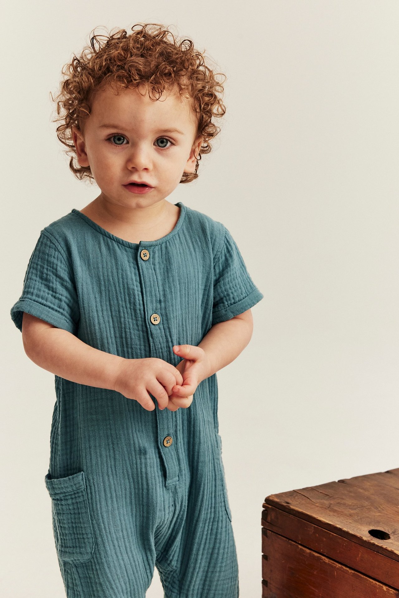 Vorderansicht eines Babys in einem dunkel türkisen Baumwoll-Musselin-Jumpsuit mit Knöpfen und aufgesetzten Taschen.