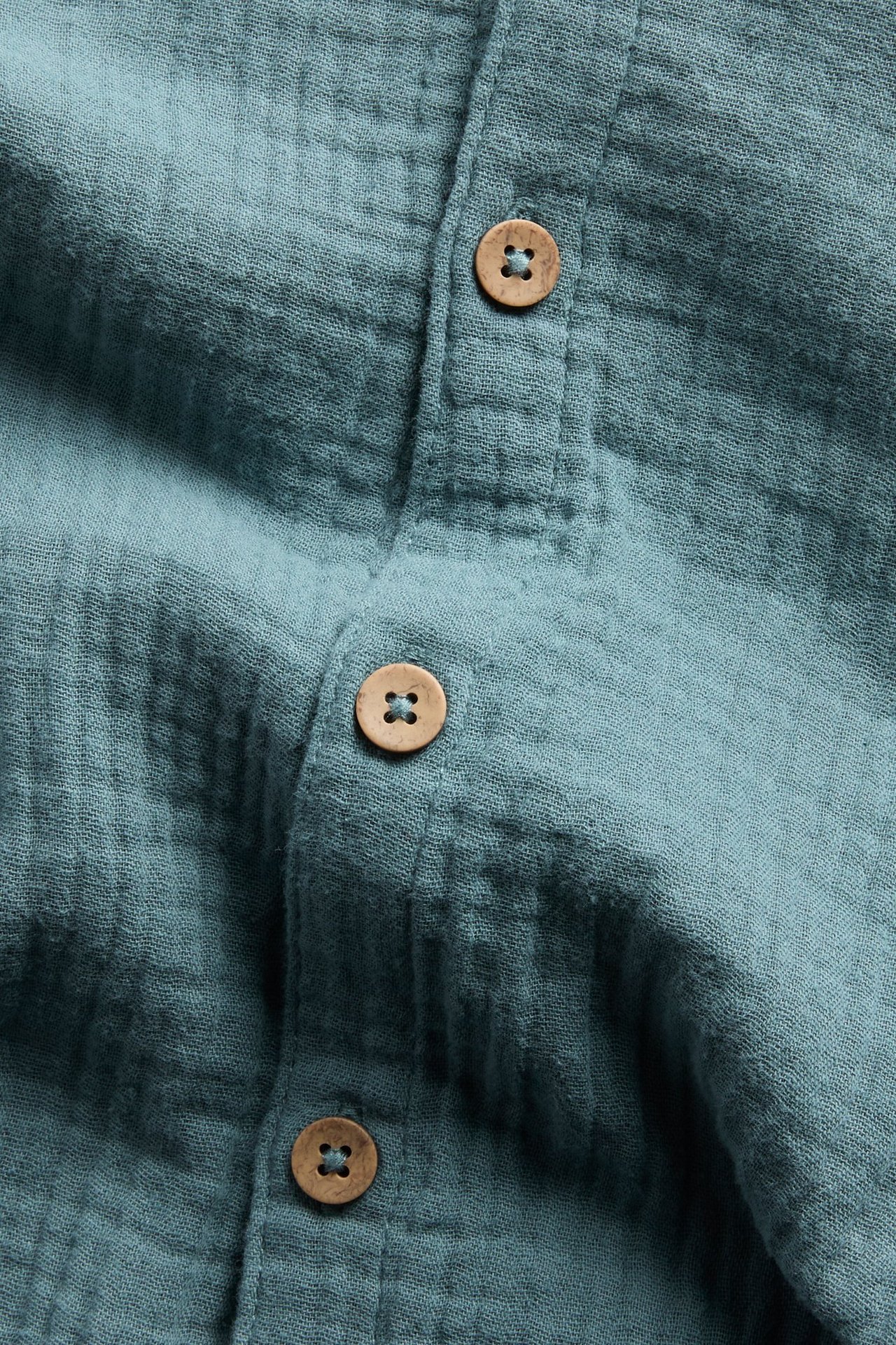 Nahaufnahme eines dunkeltürkisen kurzärmeligen Musselin-Overalls mit hellbraunen Holzknöpfen.