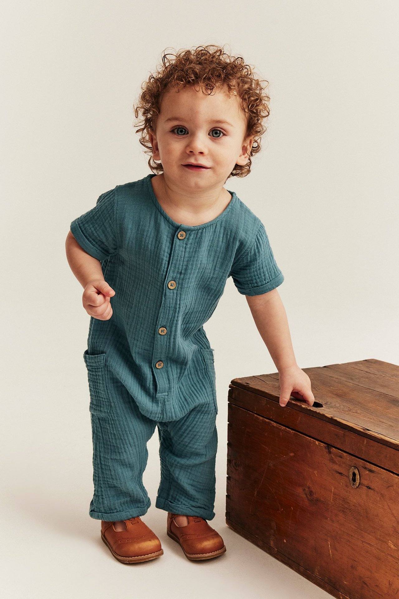 Baby trägt einen dunkel-türkisfarbenen kurzärmeligen Baumwoll-Musselin-Overall mit Knöpfen, Taschen und braunen Schuhen.