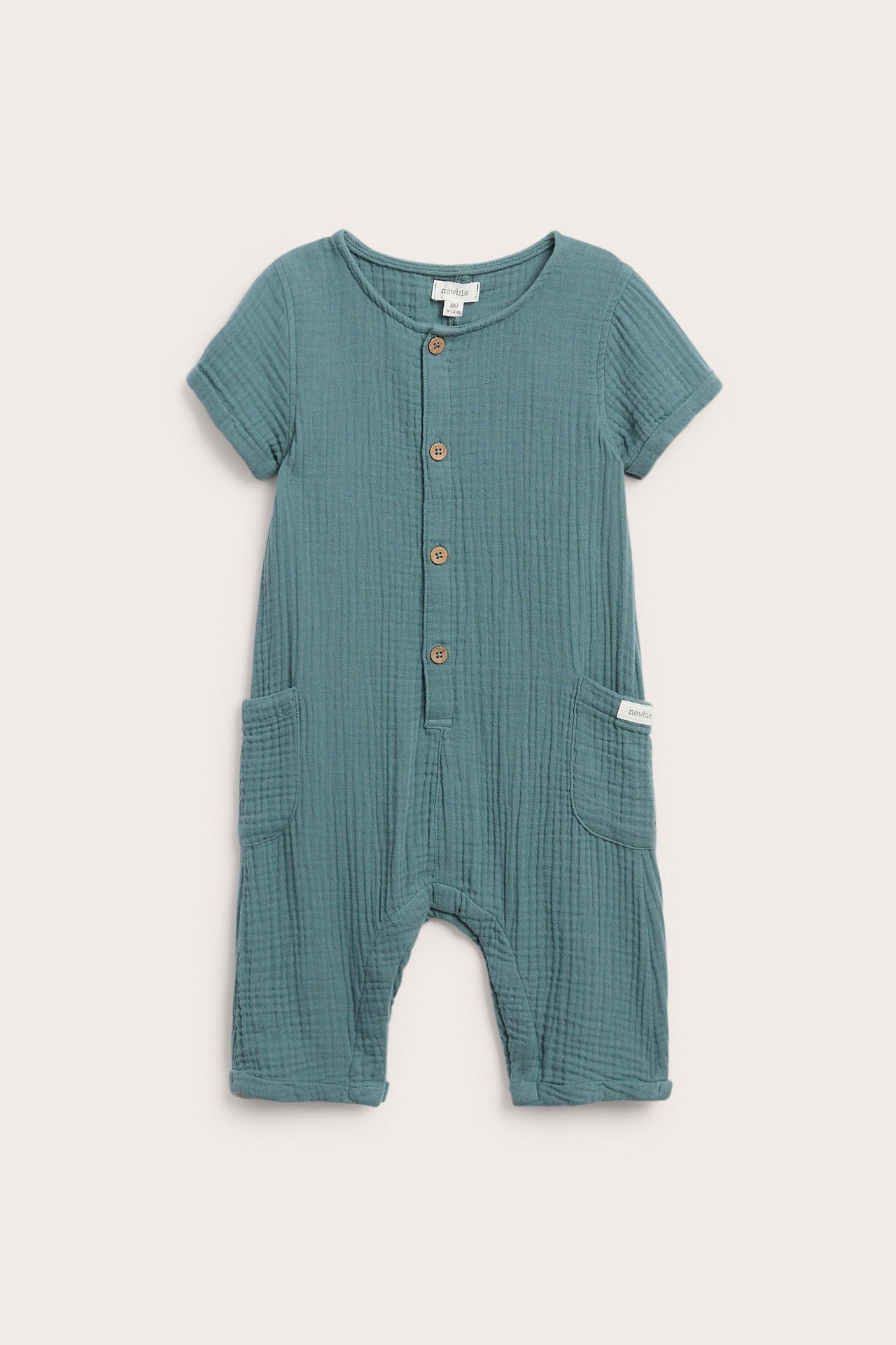 Vorderansicht eines dunkel türkisen Baby-Jumpsuits aus Baumwollmusselin mit kurzen Ärmeln, Knöpfen und Seitentaschen.