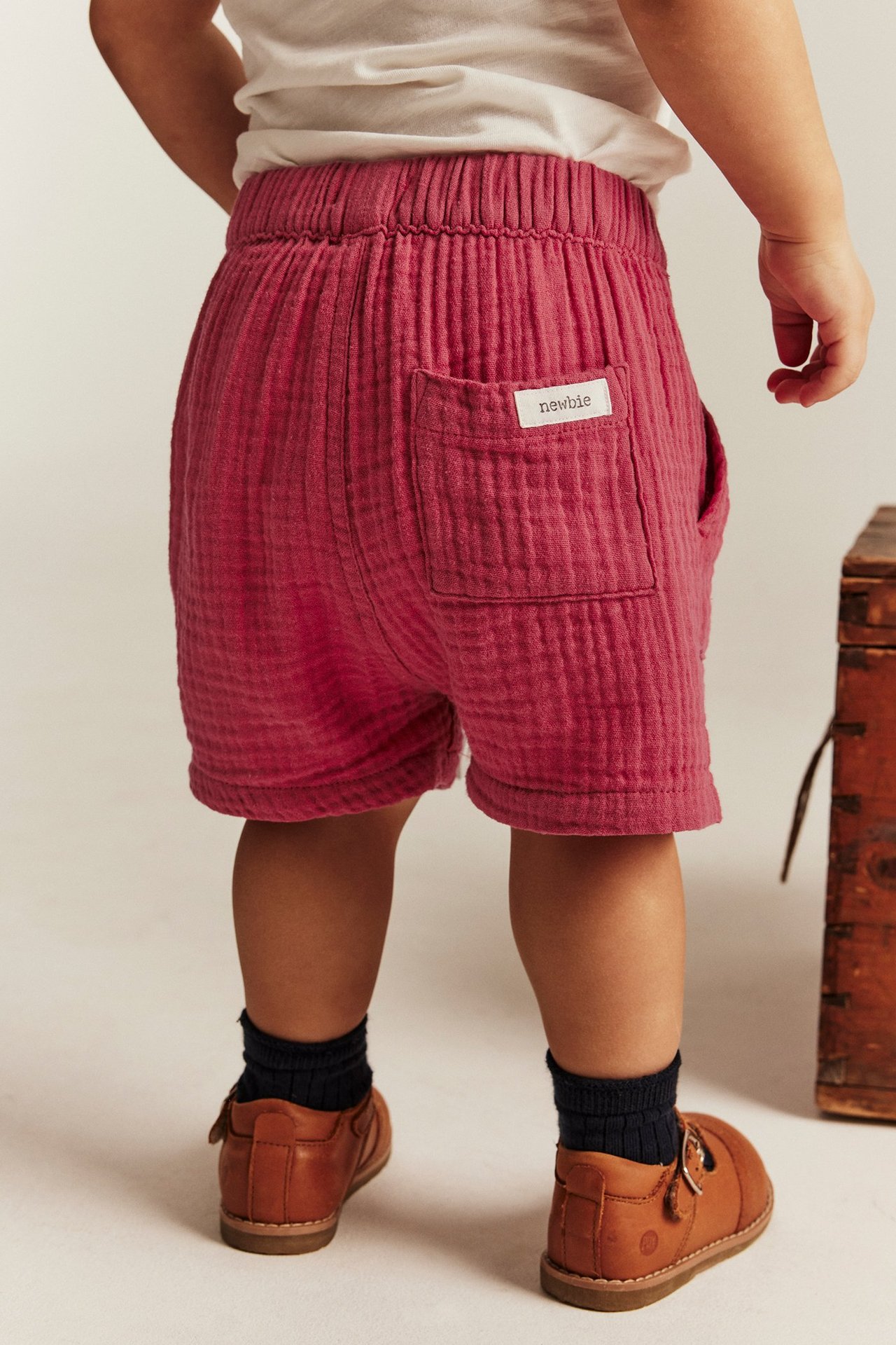 Rückenansicht Baby: rote gewebte Baumwollshorts, dunkle Socken, braune Lederschnallenschuhe.