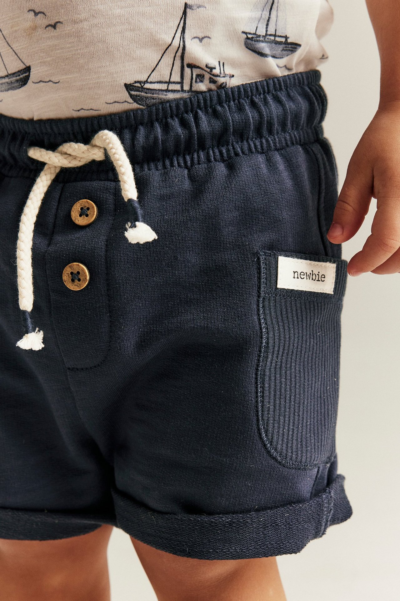 Close-up of kid's dark blue slub jersey shorts with white drawstring and buttons, worn with a boat-print t-shirt.