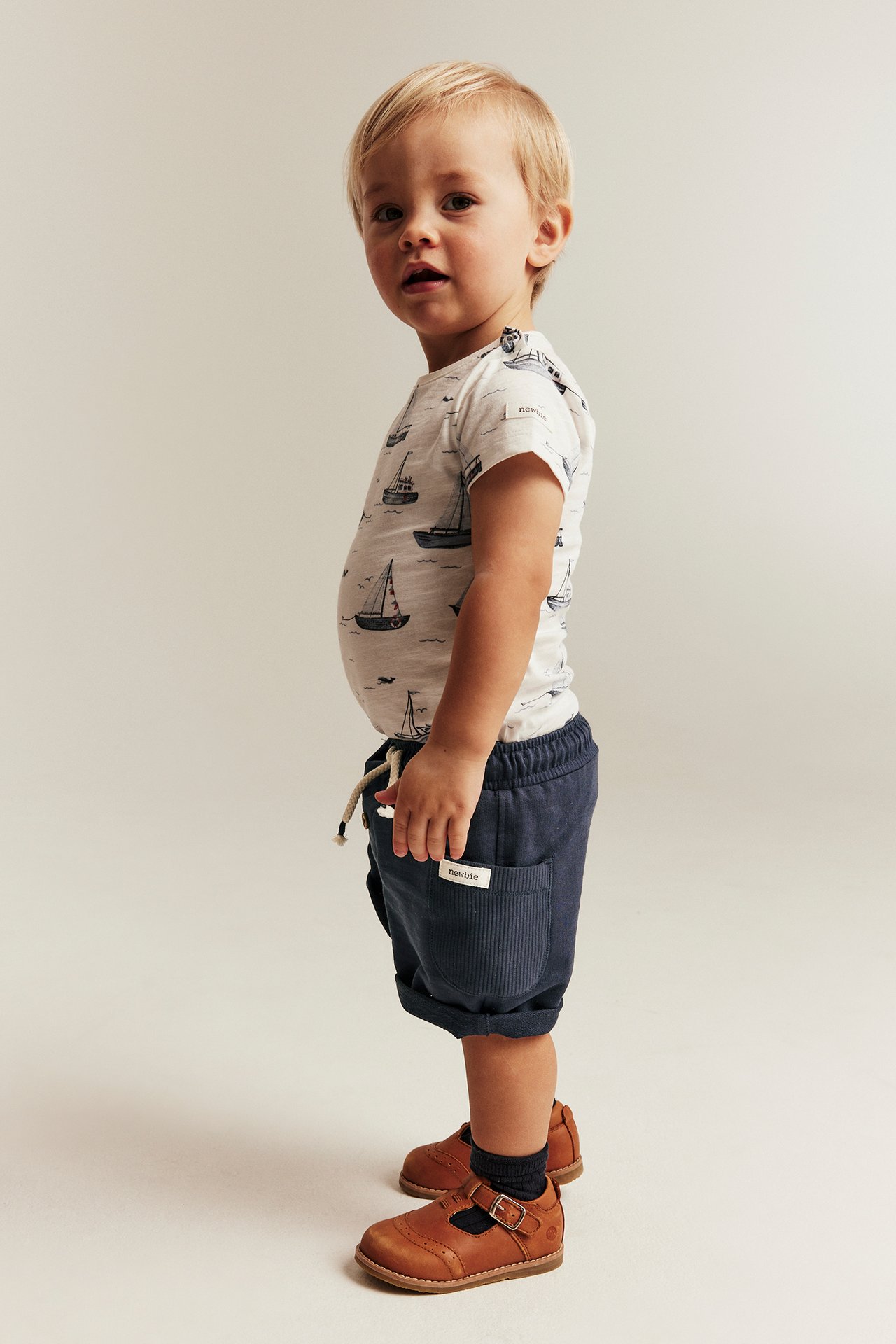 Seitenansicht eines Kindes in dunkelblauen Slub-Jersey-Shorts, weißem T-Shirt mit Bootsmuster und braunen Schuhen.