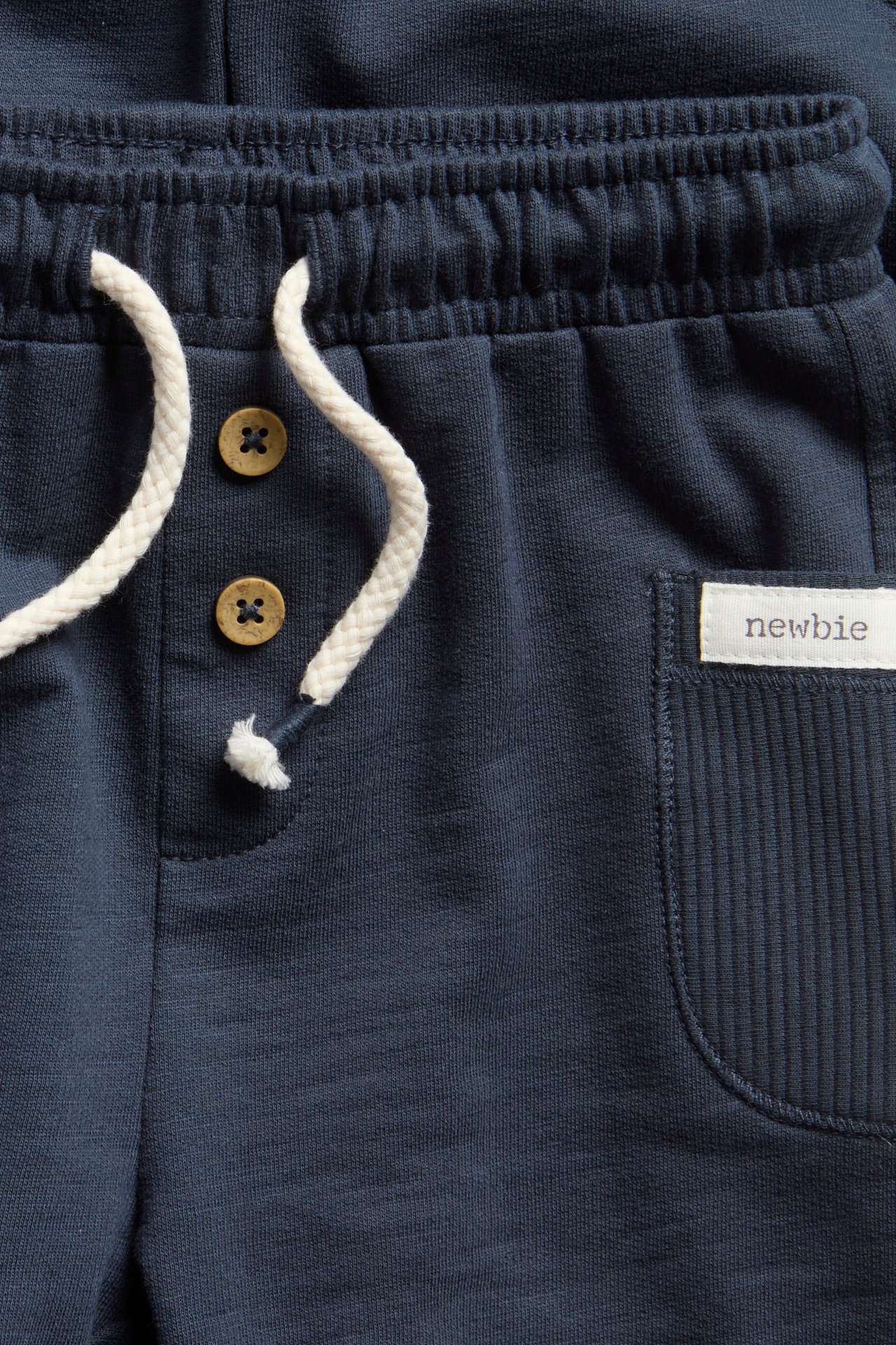Close-up: Kid's dark blue jersey shorts with elastic waist, white drawstring, brown buttons, and ribbed pocket.