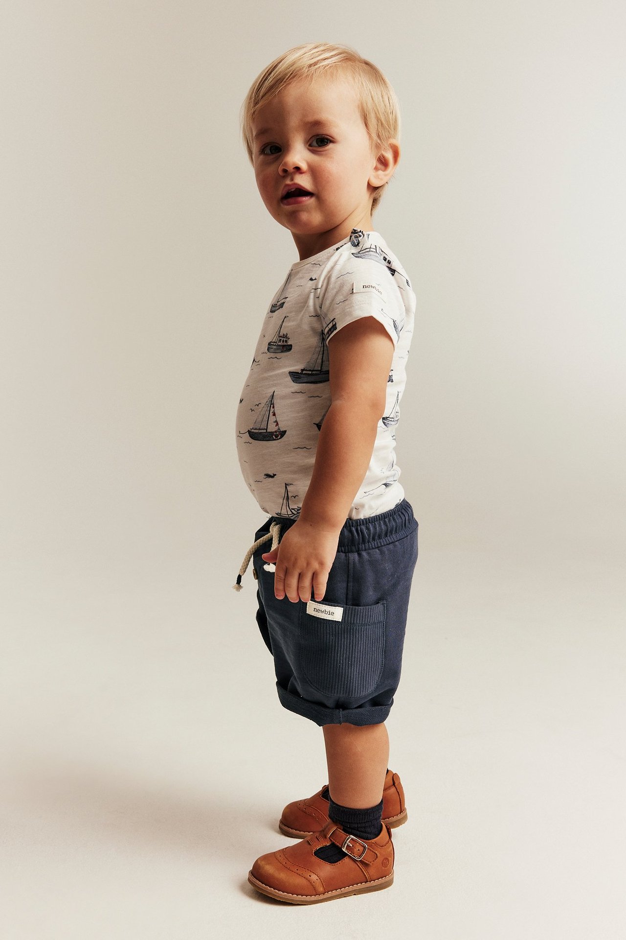 Seitenansicht: Baby trägt dunkelblaue Slub-Shorts mit Ripptaschen, umgekrempelten Säumen, ein Boot-T-Shirt und braune Schuhe.