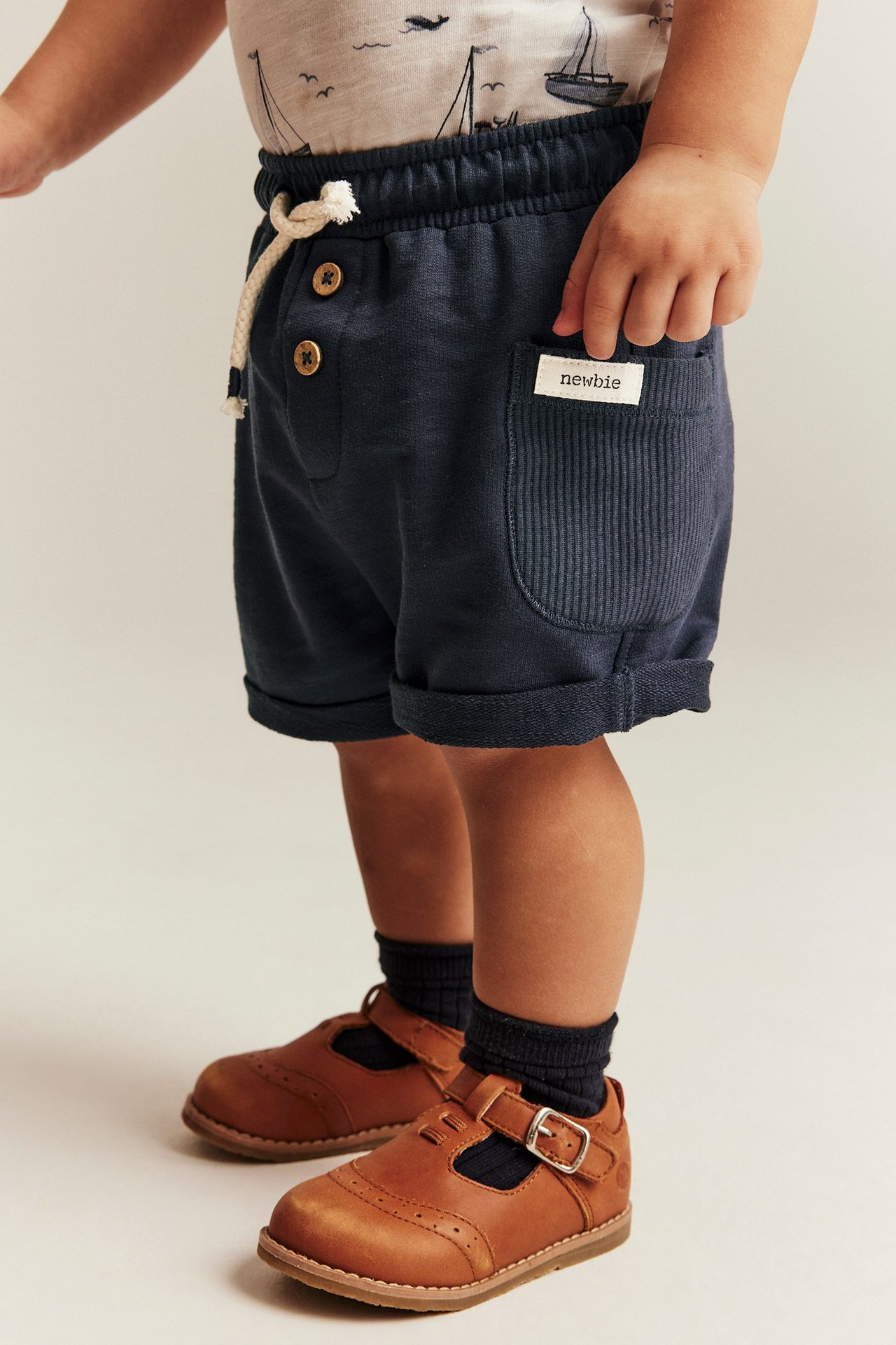 Seitenansicht Kind in dunkelblauen Slub-Jersey-Shorts mit Kordelzug, Ripptaschen und braunen Lederschuhen.