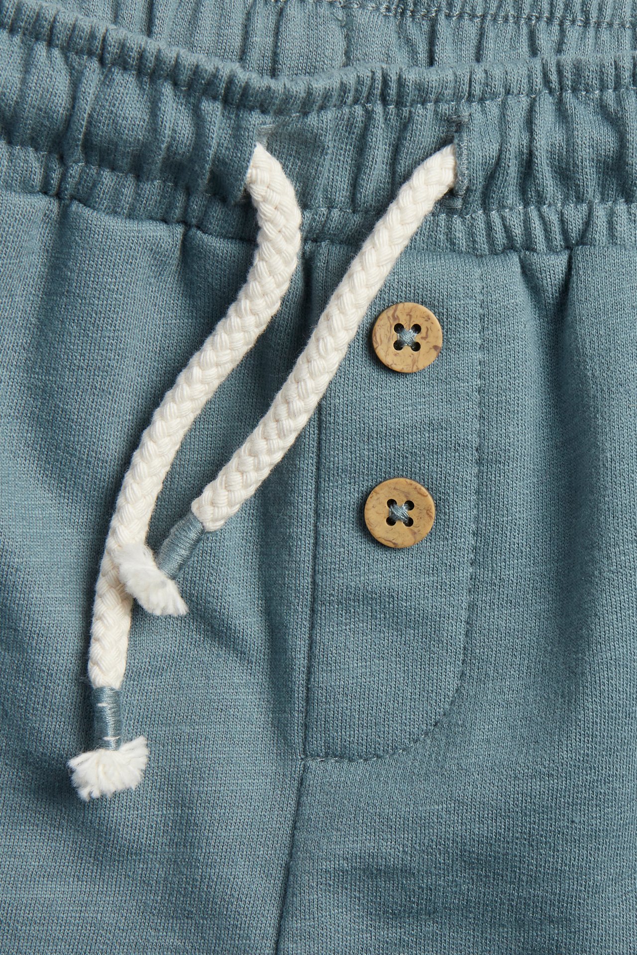 Close-up front view of dark turquoise cotton slub shorts with elastic waist, cream drawstring, and two decorative wooden buttons.