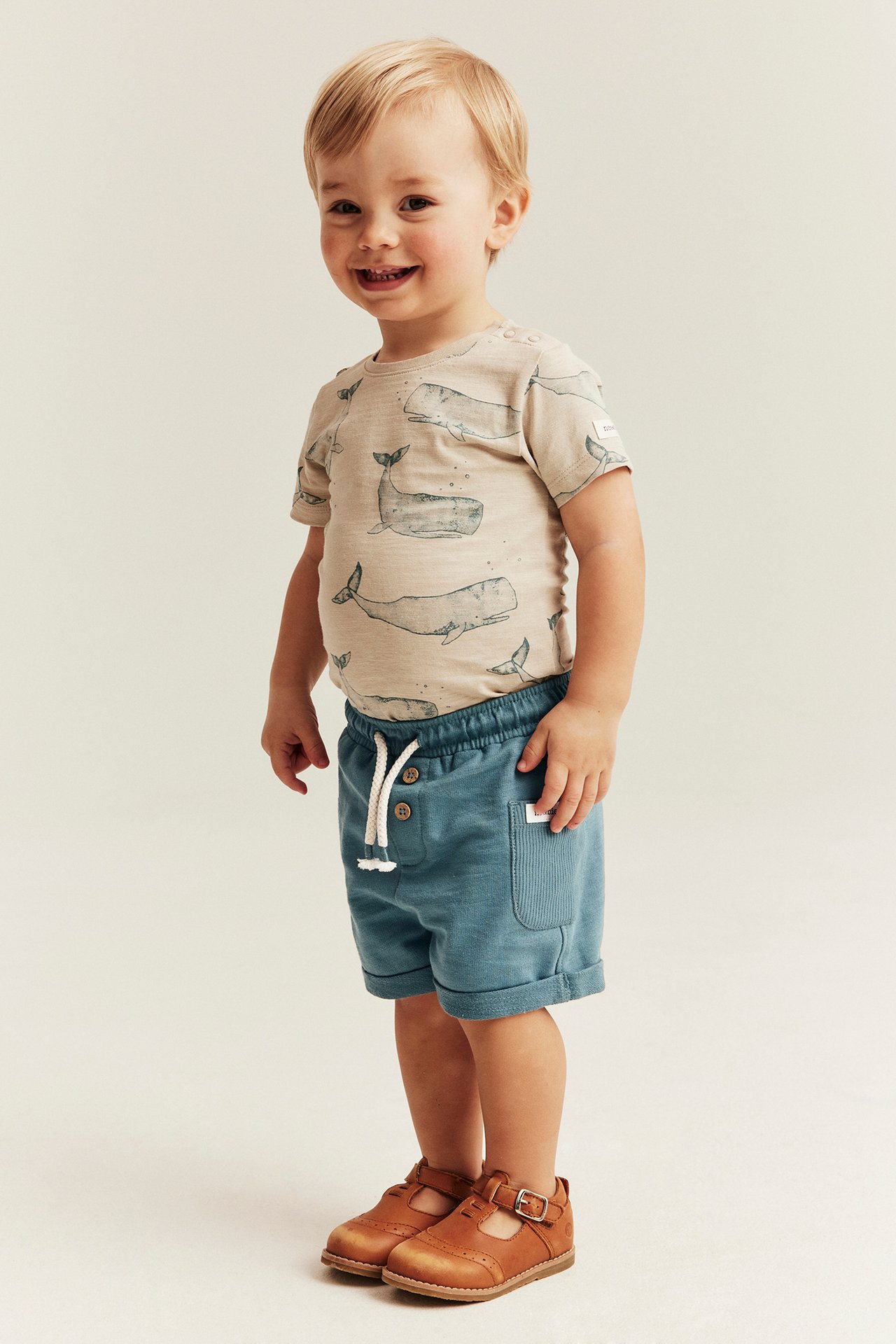 Vorderansicht, Baby in dunkeltürkisen Slub-Jersey-Shorts, Wal-T-Shirt und braunen Schuhen.