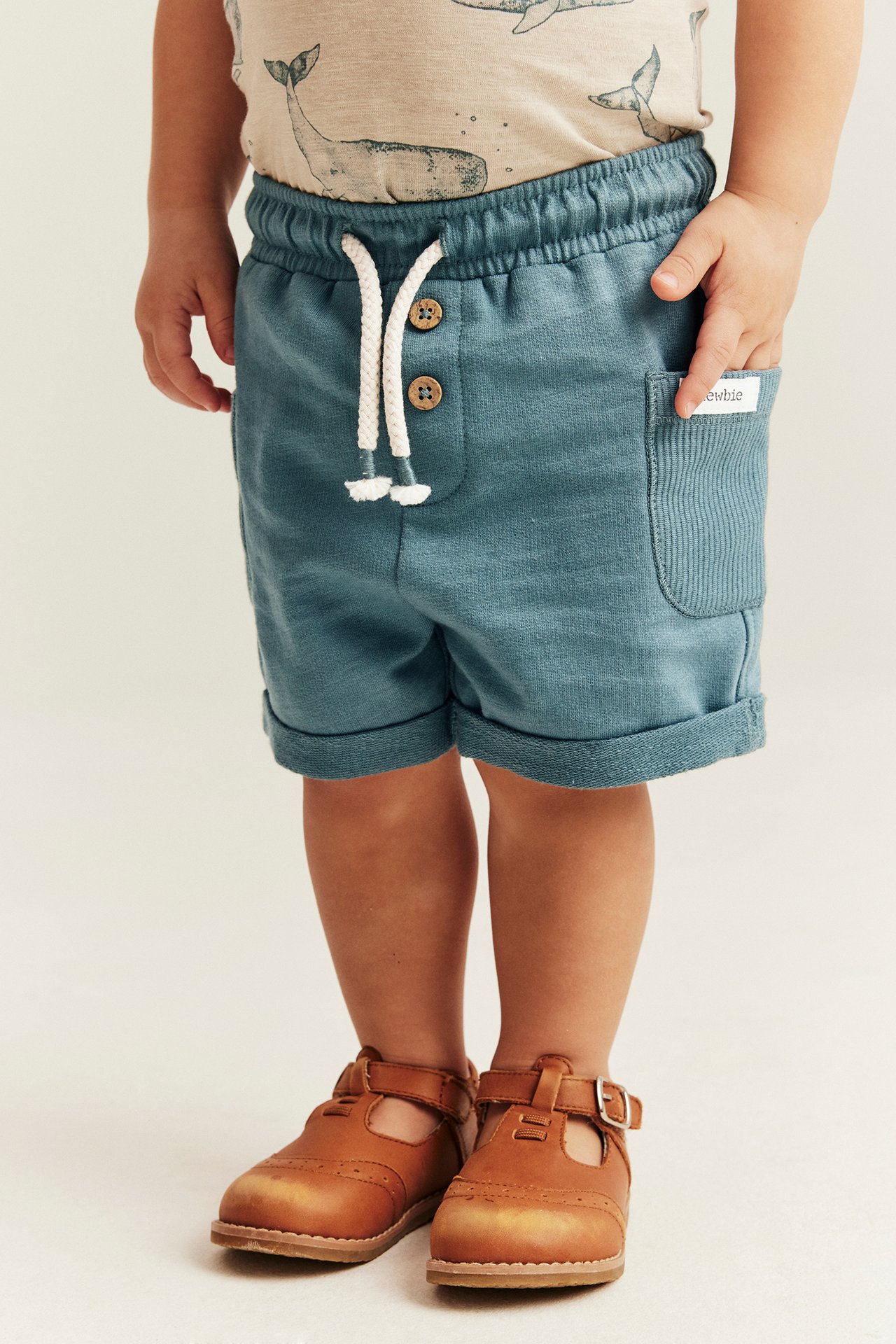Front view: Kid's dark turquoise slub shorts with drawstring and buttons. Worn with whale print t-shirt and brown shoes.