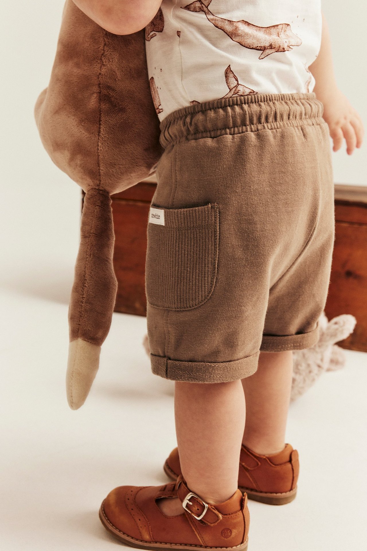 Baby trägt braune Slub-Baumwoll-Shorts, ein T-Shirt mit Walmotiv und braune Lederschuhe.
