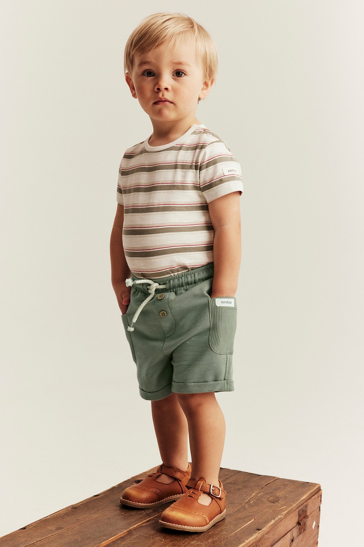 Vorderansicht eines Kindes in dunkelgrünen Slub-Jersey-Shorts, einem gestreiften T-Shirt und braunen T-Spangen-Schuhen.