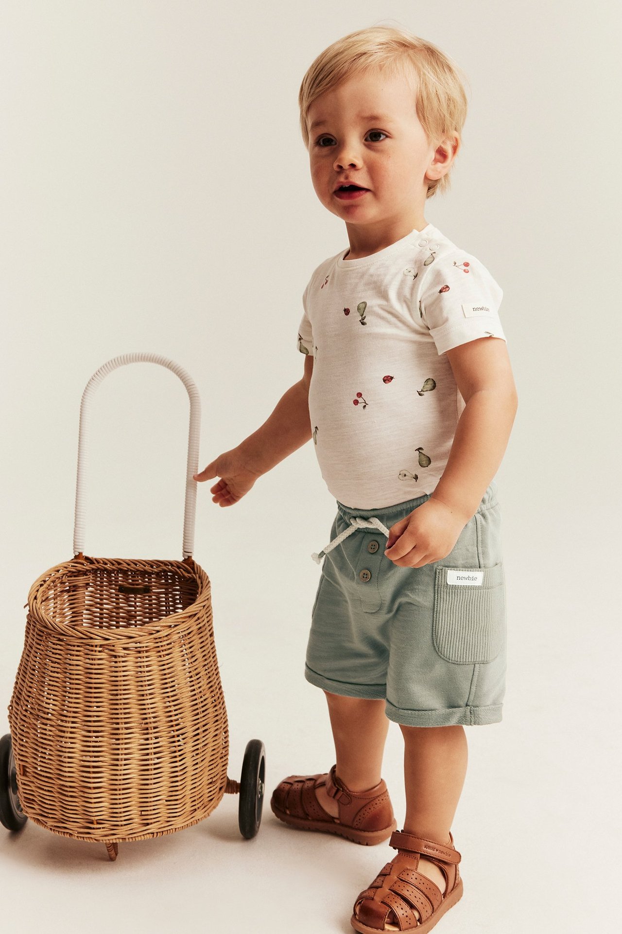 Vorderansicht: Baby in grünen Slub-Jersey-Shorts, weißem gemustertem T-Shirt und braunen Sandalen.