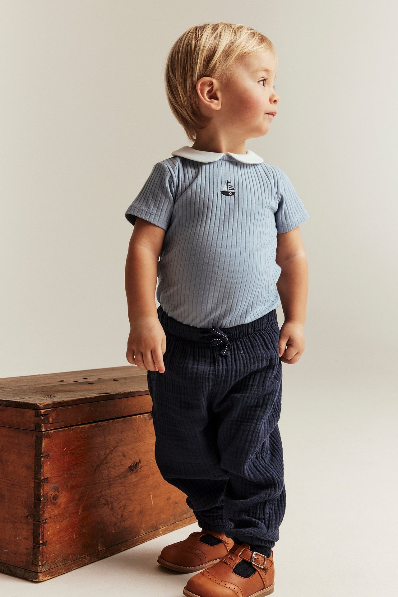 Side view, baby in light blue ribbed short-sleeved bodysuit with white collar, sailboat embroidery, and navy pants.