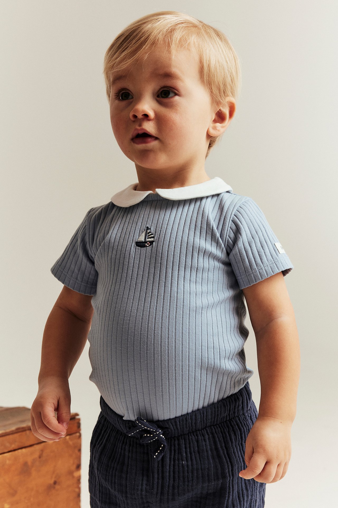 Front view: Baby wears light blue ribbed short-sleeved bodysuit with white collar, sailboat embroidery, and dark blue pants.