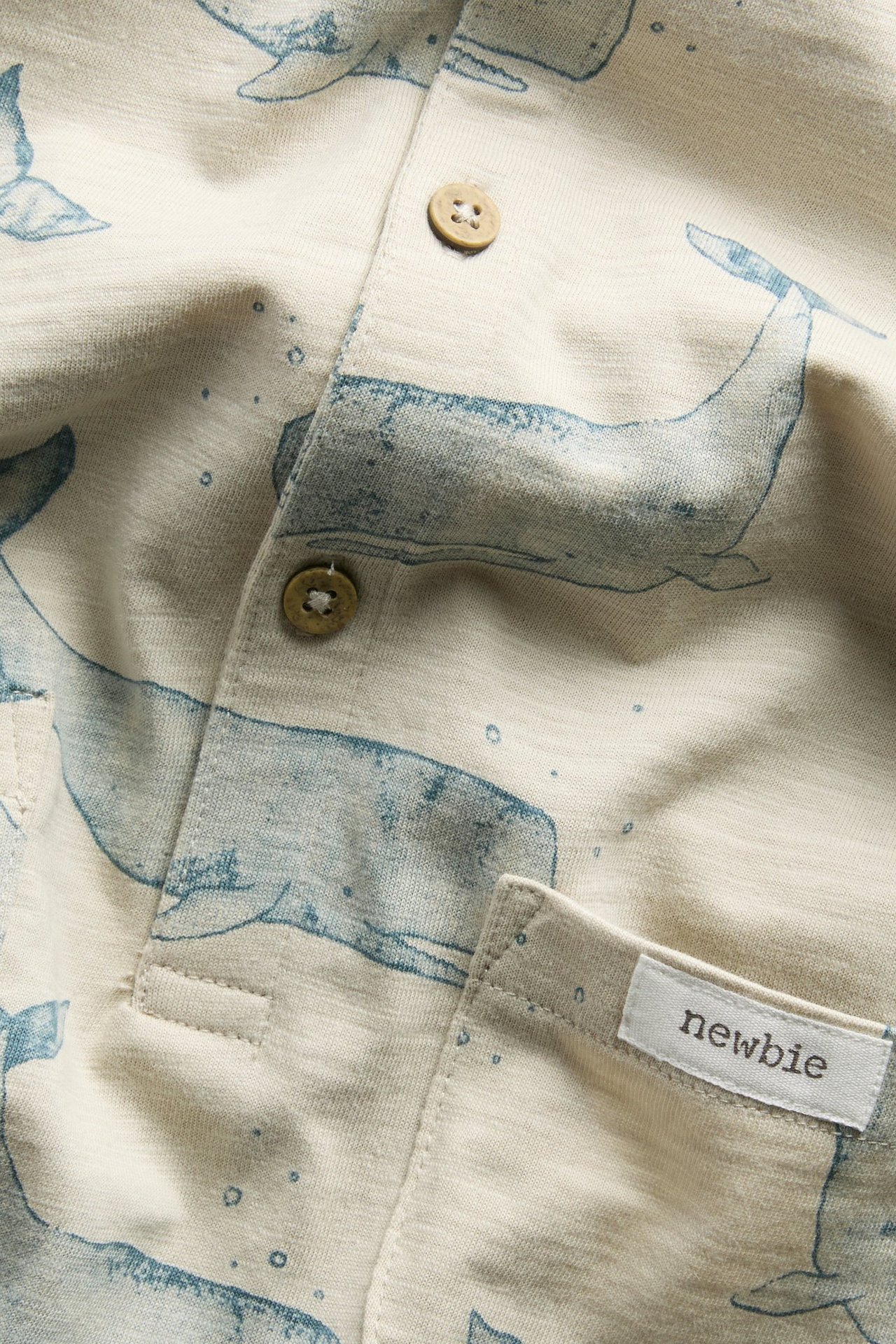 Nahaufnahme vorne eines beigen Baumwoll-Slub-Rompers mit blauem Walmuster und braunen Knöpfen.