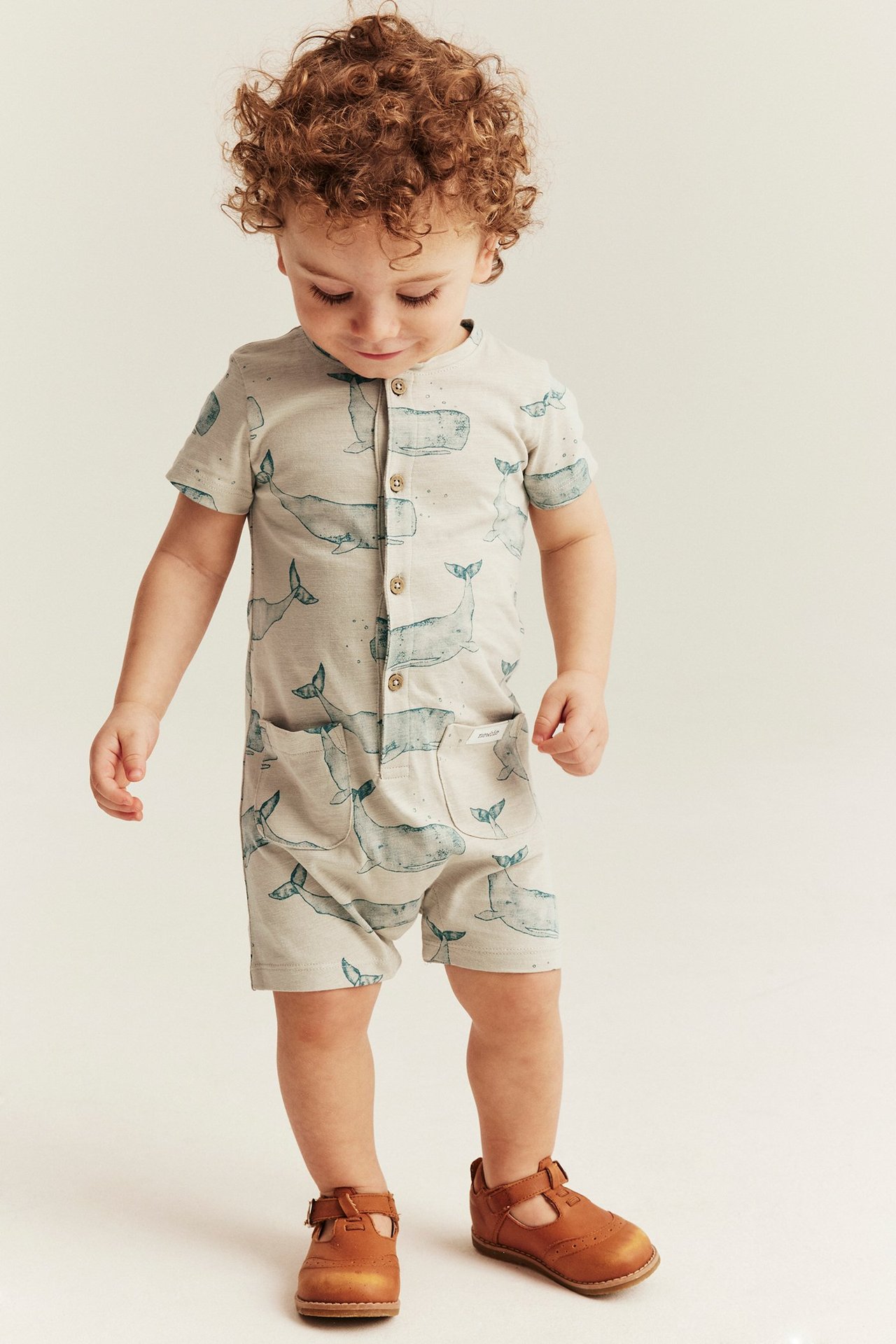 Front view: Baby in a beige cotton whale-patterned short-sleeved romper with buttons, pockets, and brown shoes.