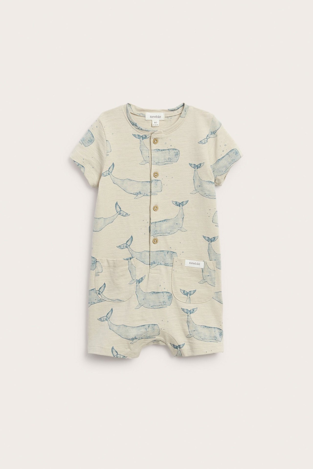 Front view of a baby's beige cotton romper with a blue whale pattern, button placket, and front pockets.