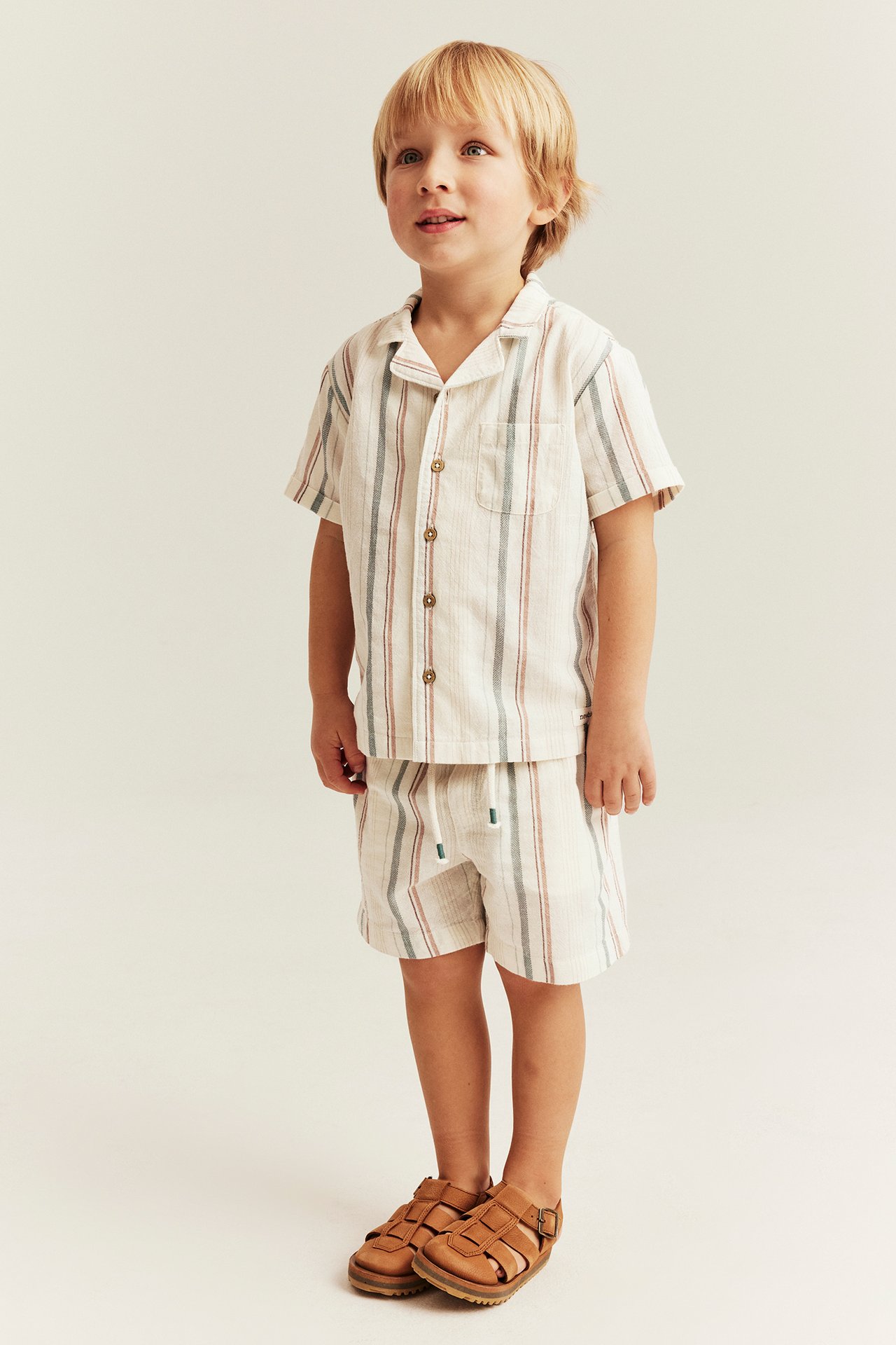 Front view of a kid in an off-white striped short-sleeved cotton shirt, matching striped shorts, and brown sandals.