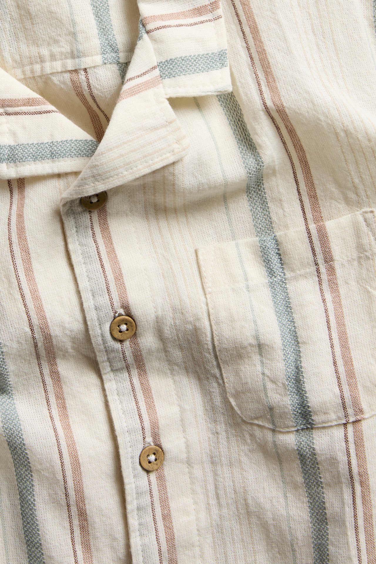 Close-up of an off-white striped short-sleeved woven shirt with a collar, wooden buttons, and a chest pocket.