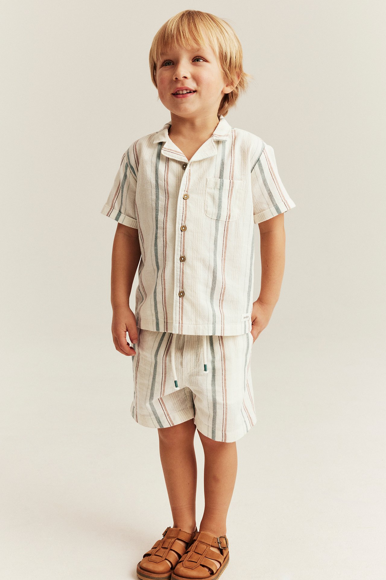 Front view: Kid wears offwhite striped cotton shorts, matching striped shirt, brown sandals.