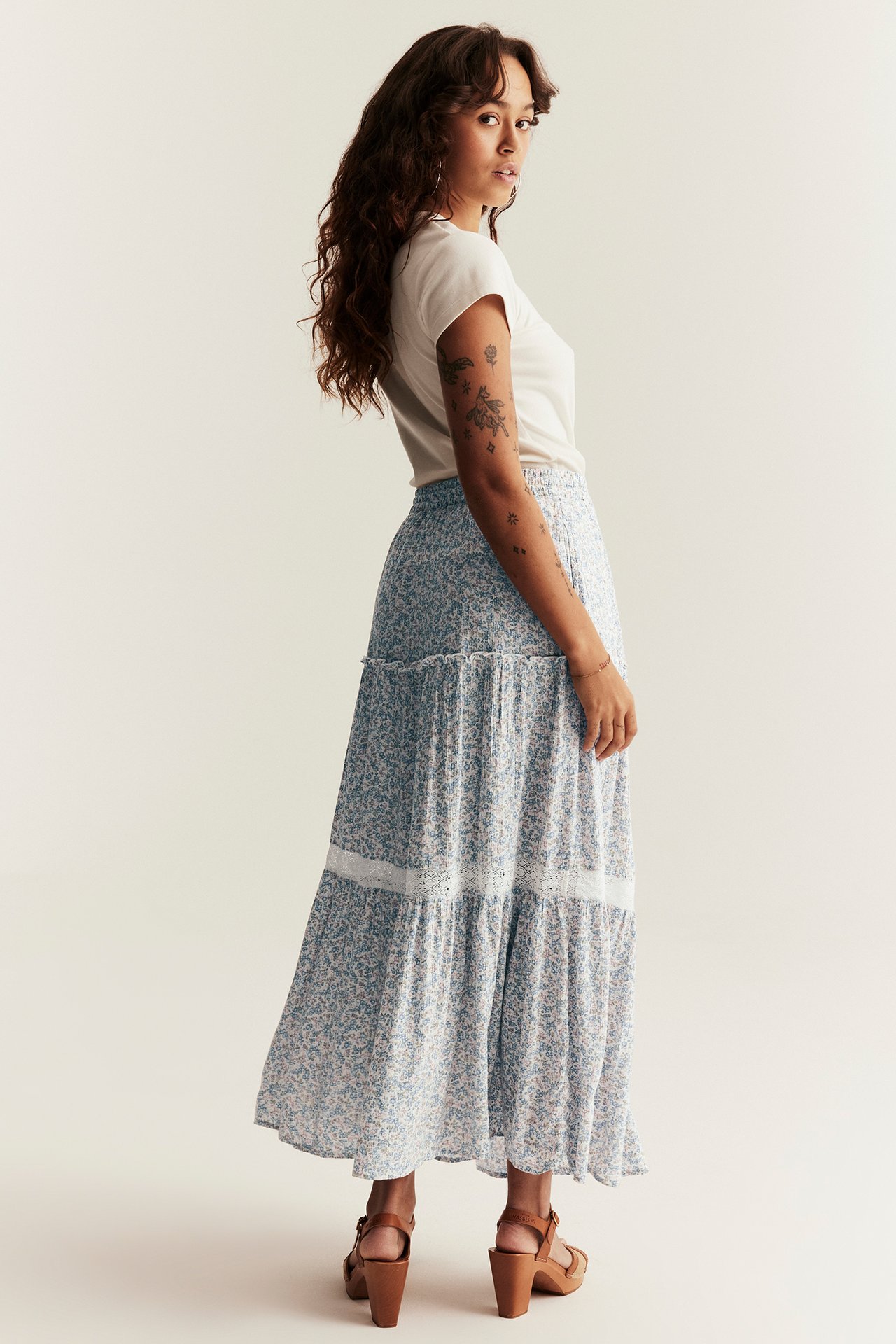 Woman in a light blue floral tiered long skirt with white lace, off-white t-shirt, and brown sandals.