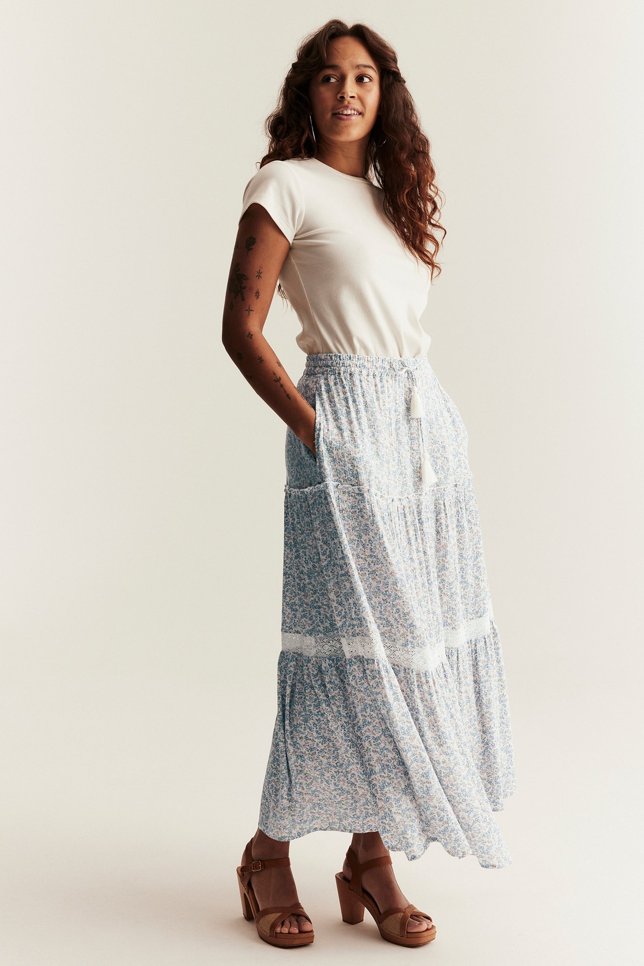 Vorderansicht: Frau in hellblauem, geblümtem Viskose-Maxirock mit Spitze, einem cremefarbenen T-Shirt und braunen Sandalen.