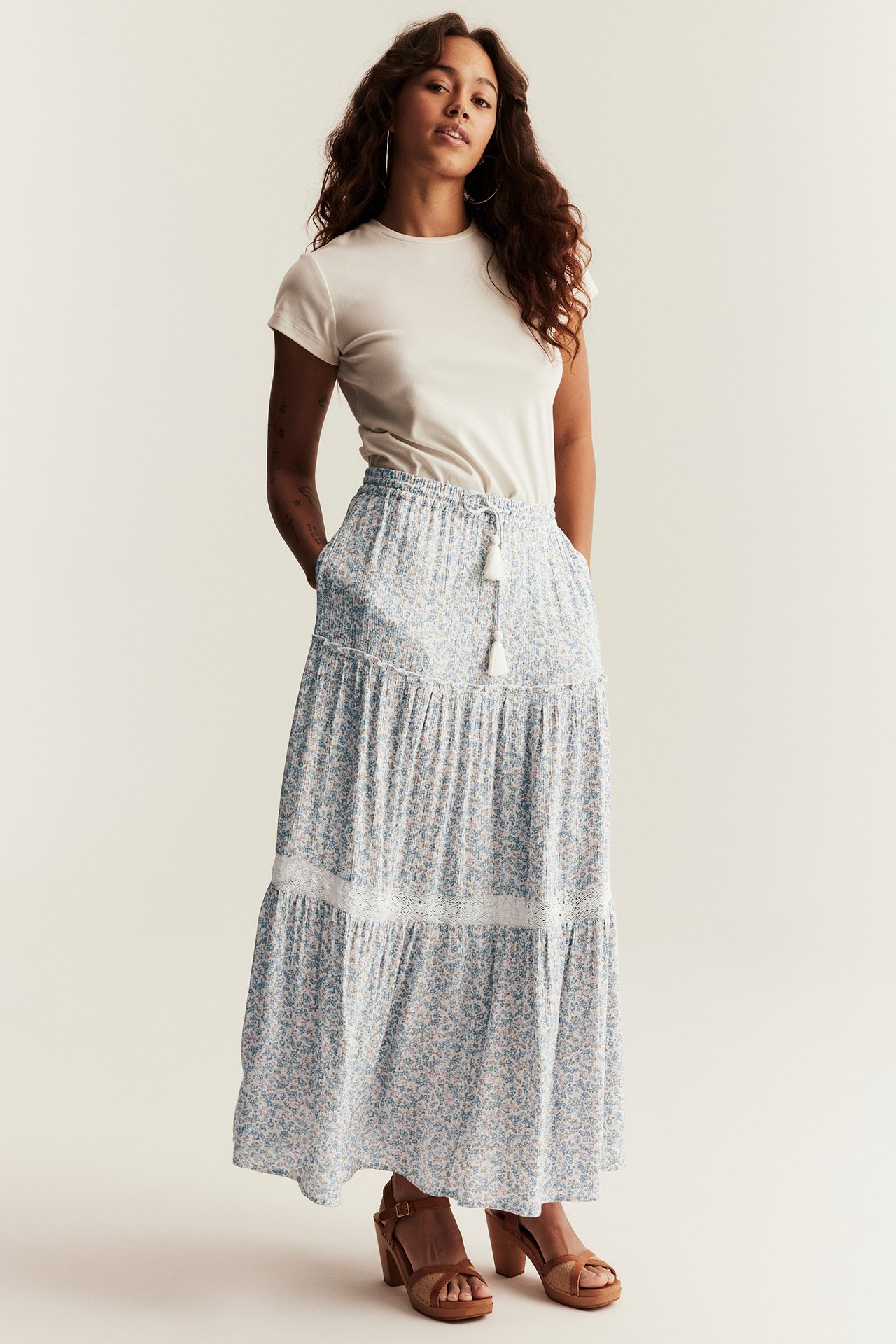 Front view: Woman in off-white and light blue floral long tiered skirt, an off-white tee, and brown sandals.