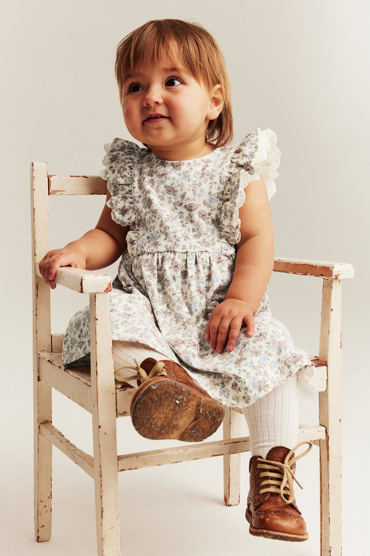 Front view of a baby wearing an off-white sleeveless floral cotton dress with ruffle lace trim, white tights, and brown shoes.