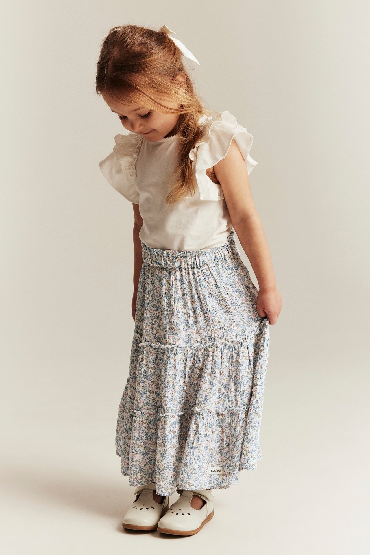 Front view: Kid wears an off-white tiered floral long skirt, white ruffled t-shirt, and white shoes.
