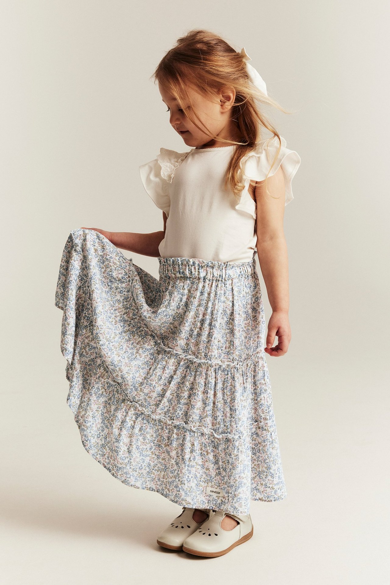Front view of a kid wearing an off-white floral tiered long skirt, a white ruffled-sleeve top, and white Mary Jane shoes.