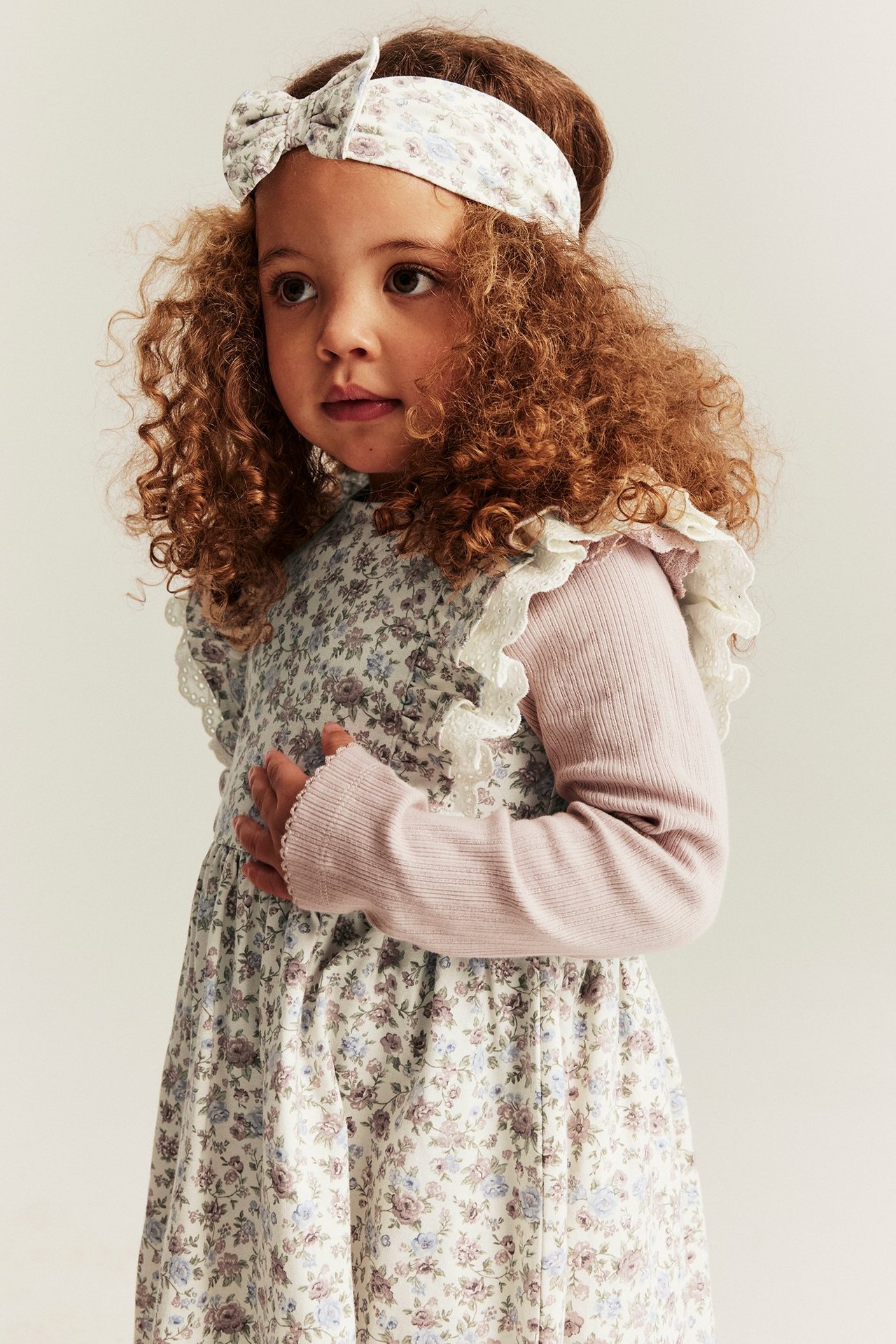 Close-up front view of a kid in an off-white floral sleeveless dress with lace, over a pink top and matching headband.