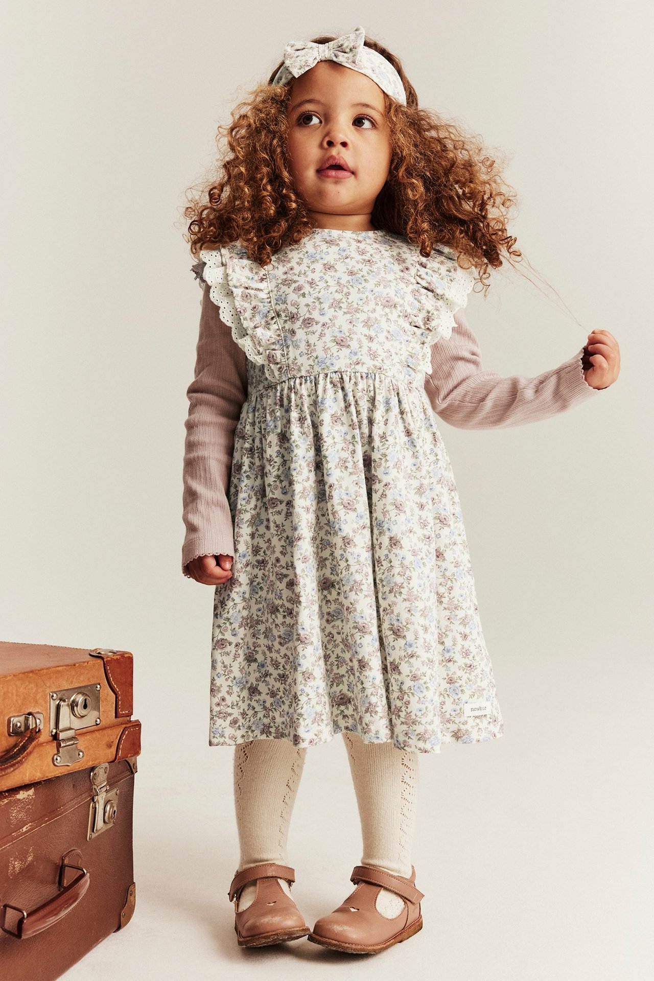 Front view: Kid in offwhite floral dress with ruffles, pink shirt, matching headband, beige tights, and brown shoes.