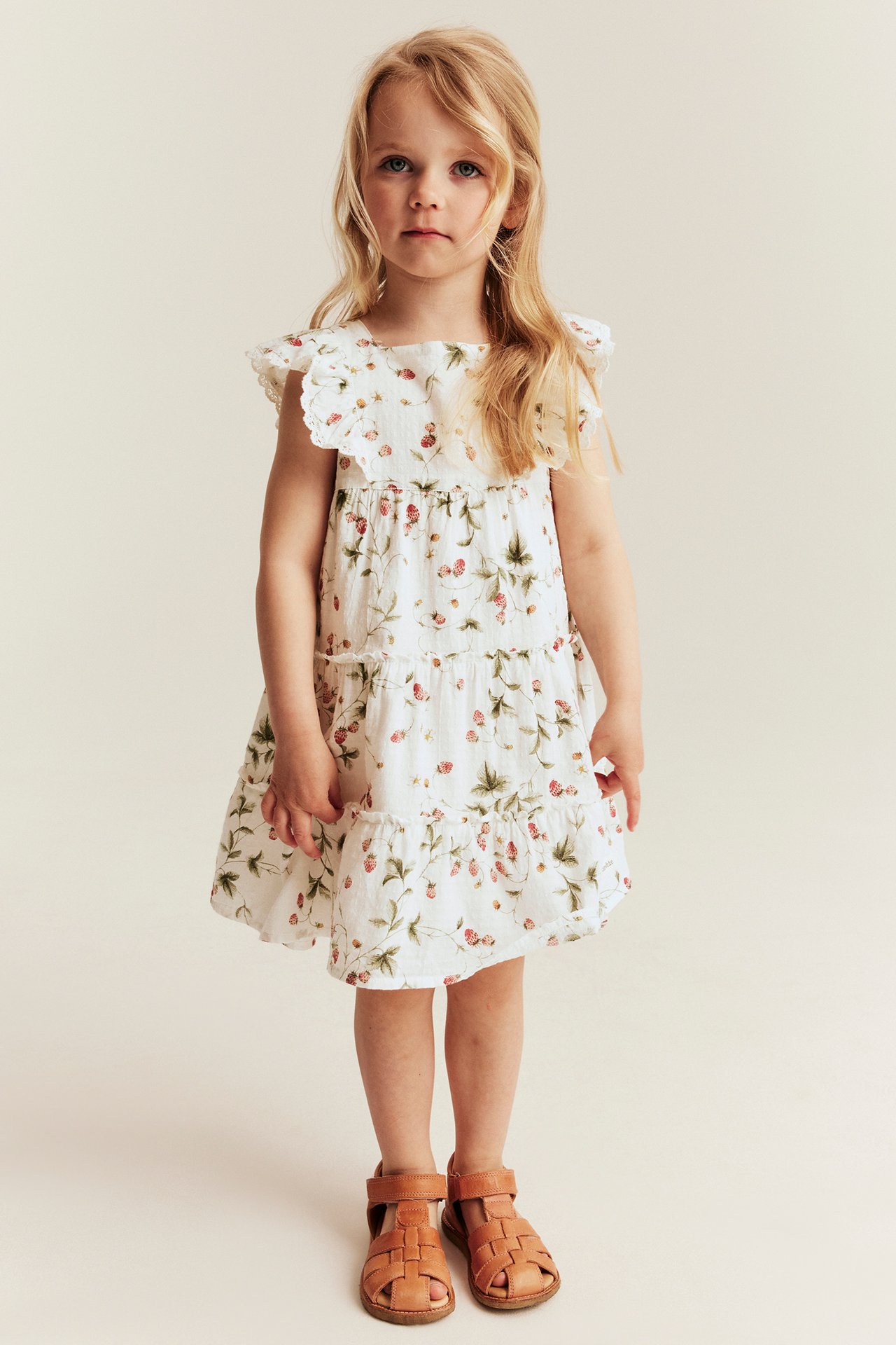 Vorderansicht, ein Mädchen trägt ein cremeweißes gemustertes gewebtes Kleid mit Rüschen und braune Ledersandalen.
