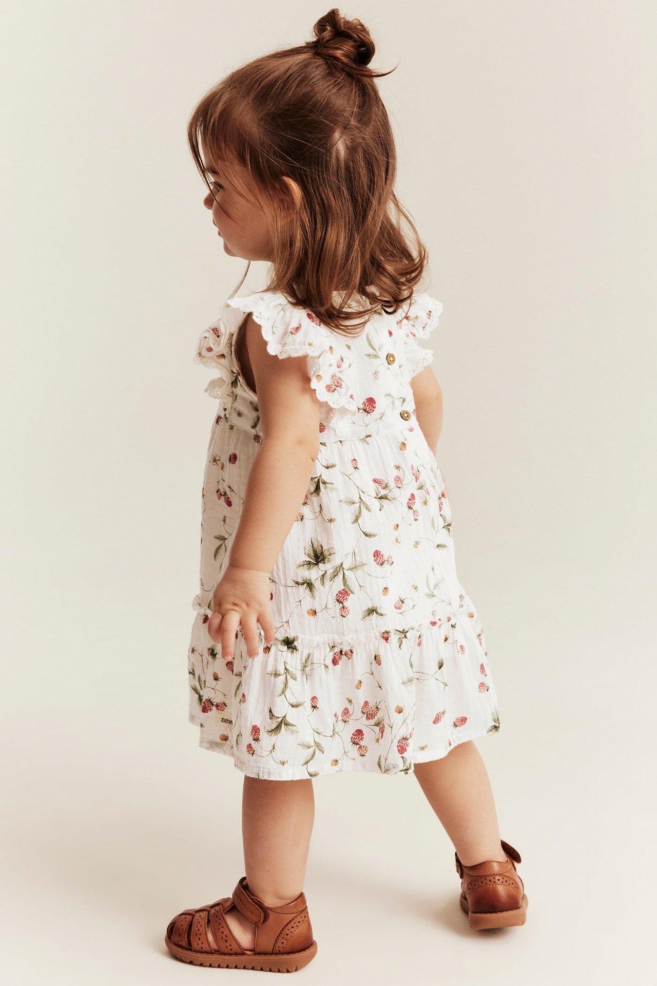 Baby-Mädchen in einem offwhite gewebten Erdbeer-Kleid mit Rüschen, Spitze, Rückenknöpfen und braunen Sandalen.