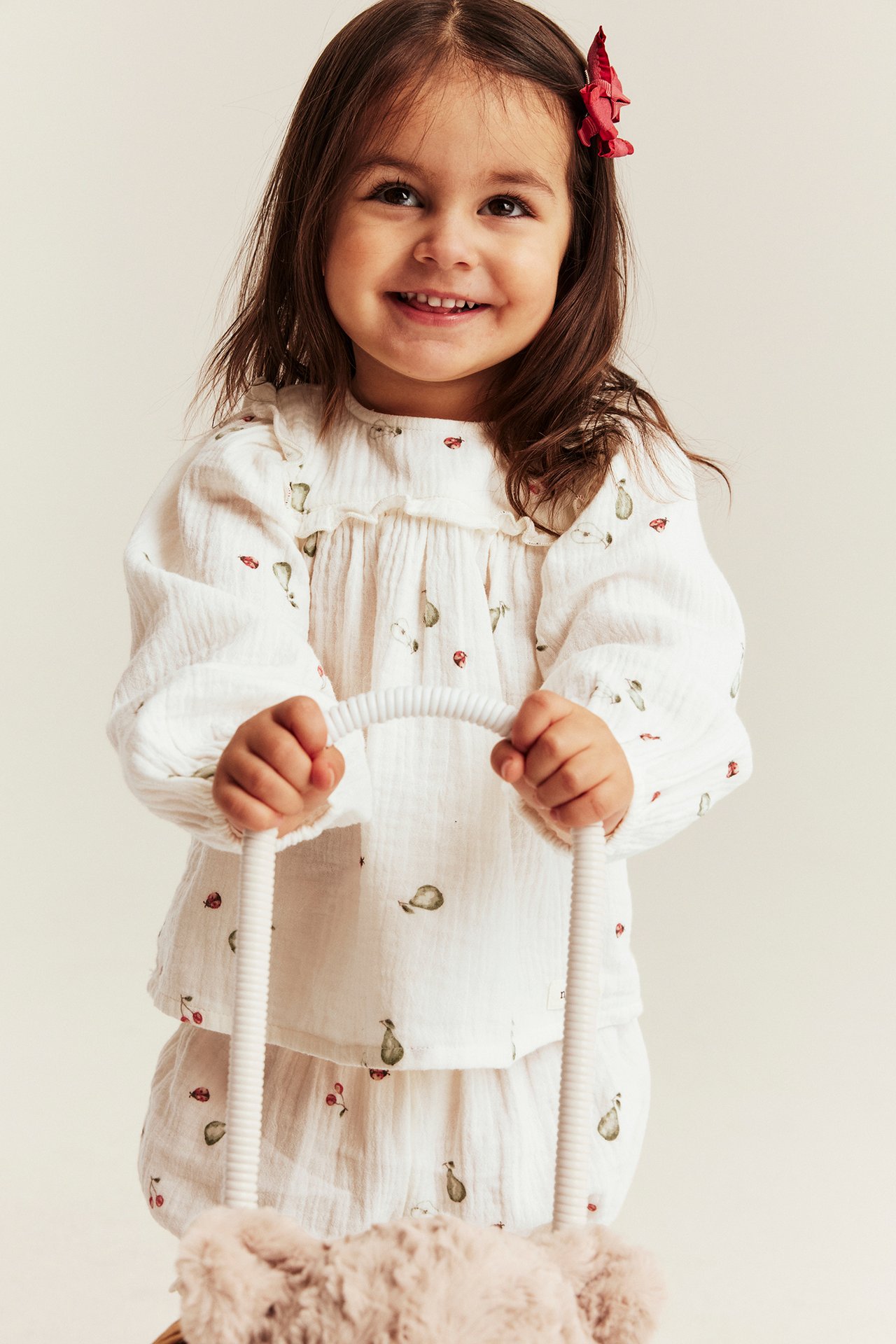 Front view: Kid in off-white patterned muslin blouse and matching shorts, holding a fuzzy toy.