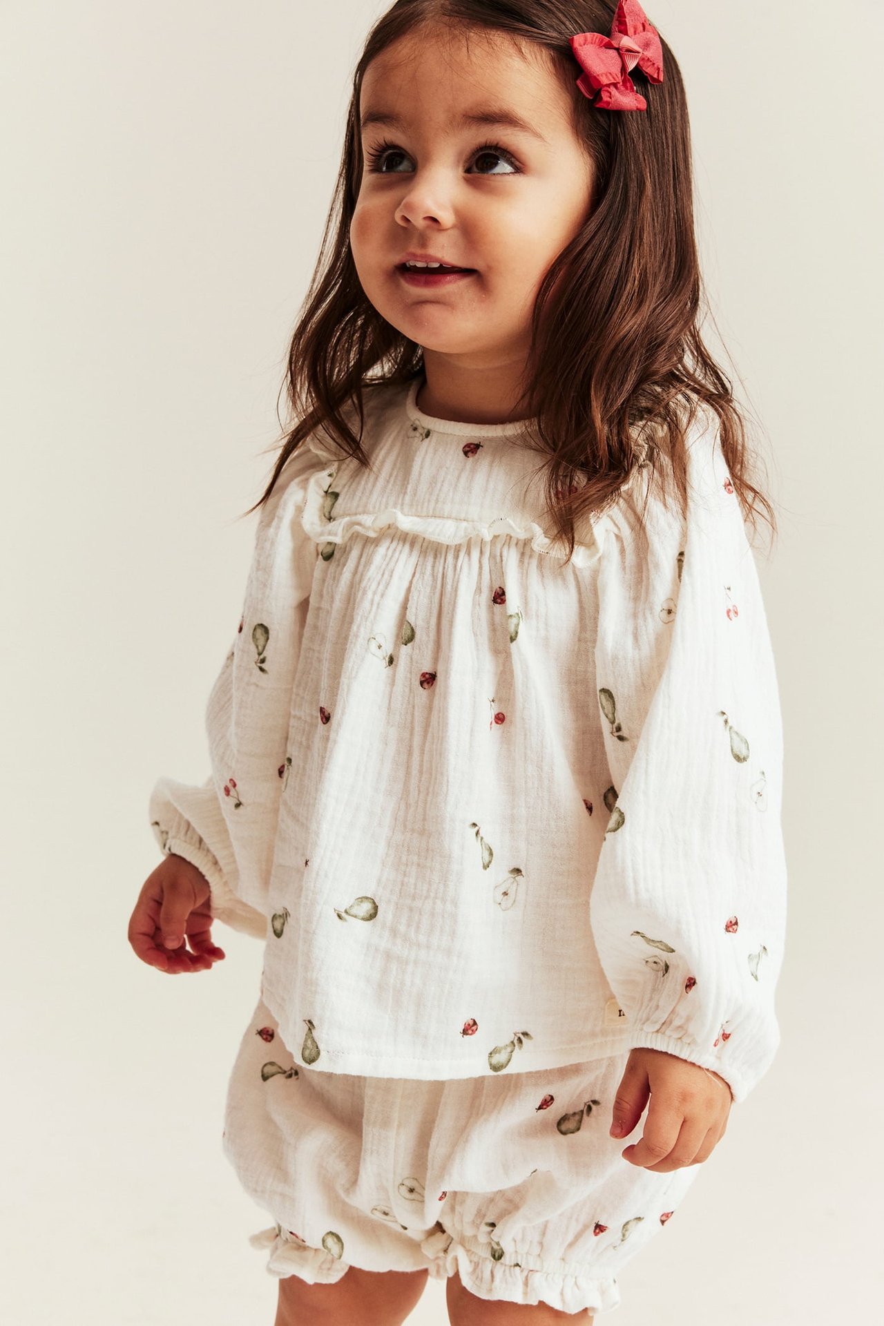 Front view of baby girl in off-white patterned muslin set with ruffled blouse, matching shorts, and red bow.